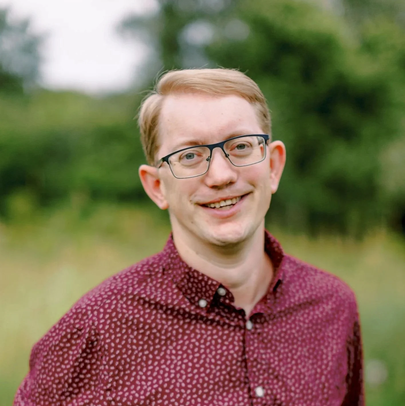 A smiling man with red hair, glasses, and wearing a maroon button-up shirt with small white polka dots, standing outdoors with blurred green trees in the background.