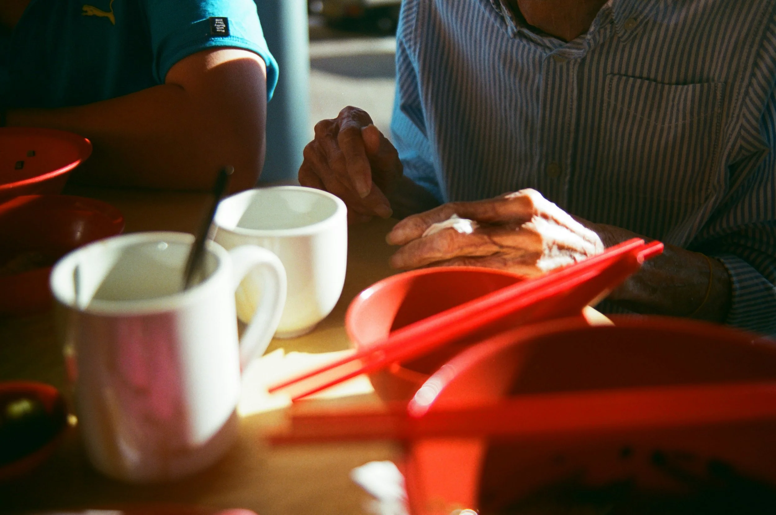 Close-up of two people holding hands across a table, with cups of coffee or tea and red trays in the foreground.