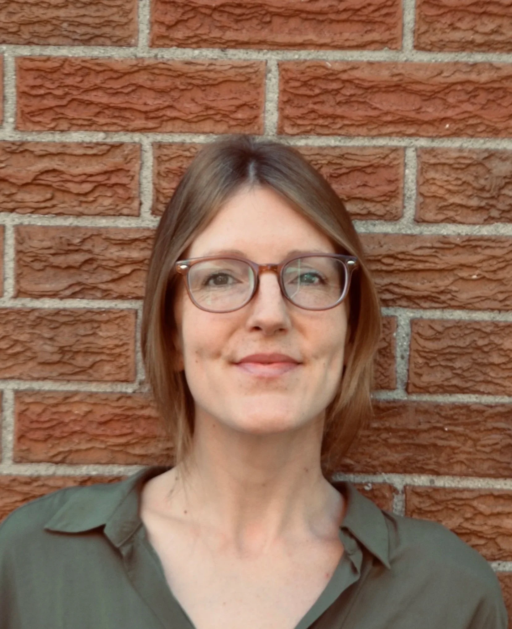 A woman with shoulder-length brown hair, glasses, and a slight smile is standing in front of a red brick wall, wearing a dark green shirt.
