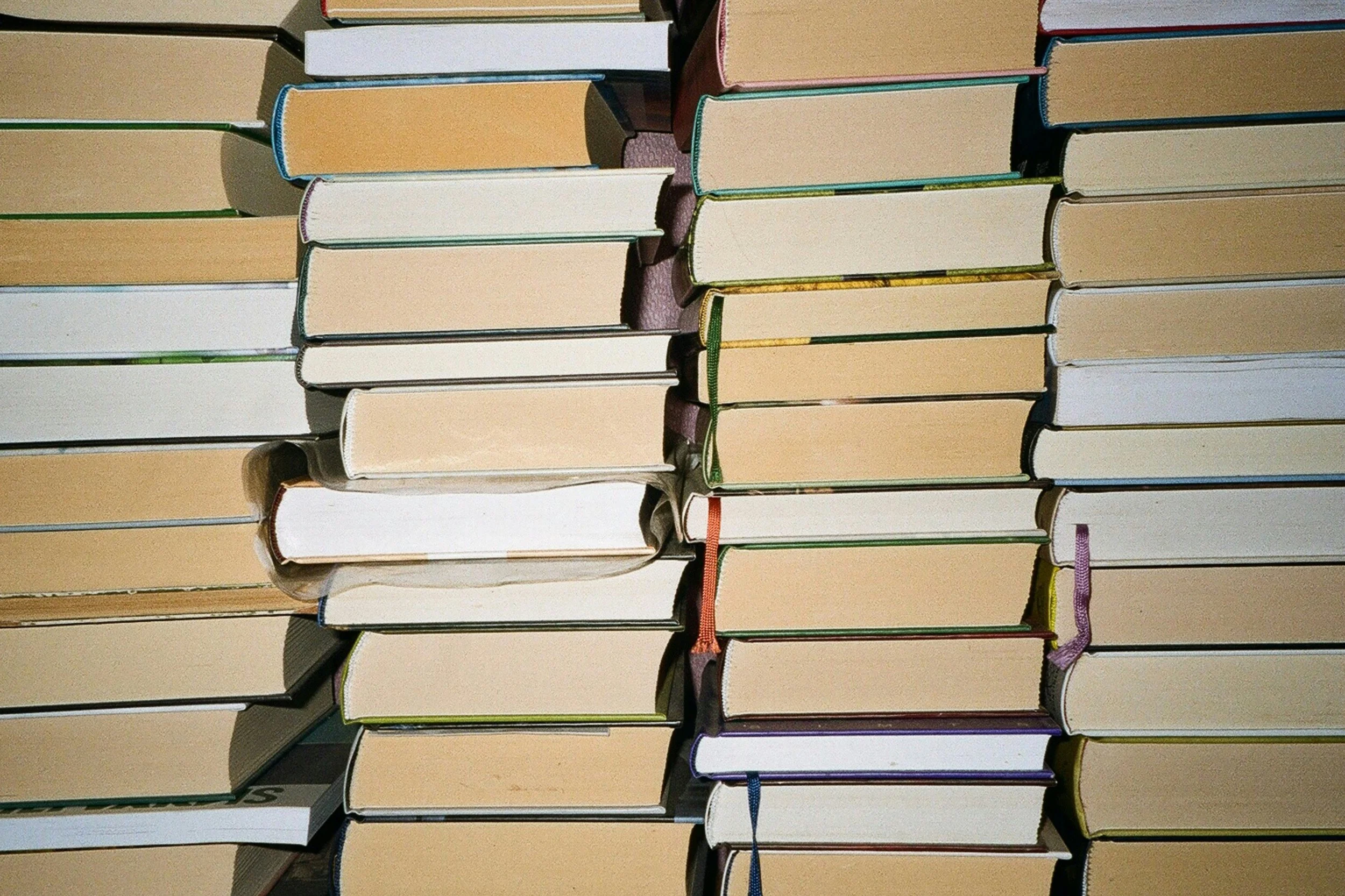A collection of books stacked horizontally with their covers facing downward, showing the spines and bookmarks in varying colors.