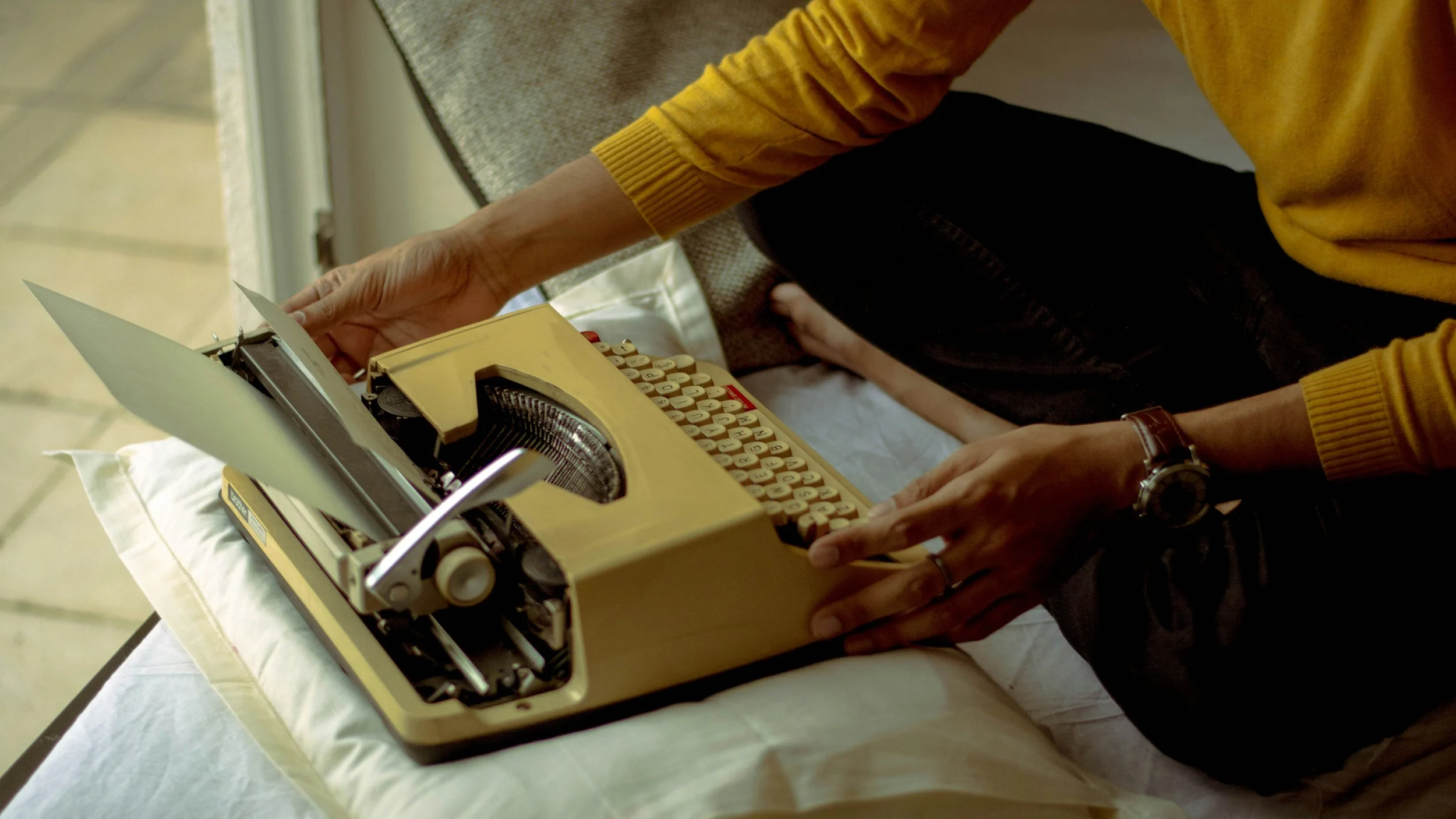 Person in a yellow sweater typing on a vintage yellow typewriter placed on a white cloth-covered table.