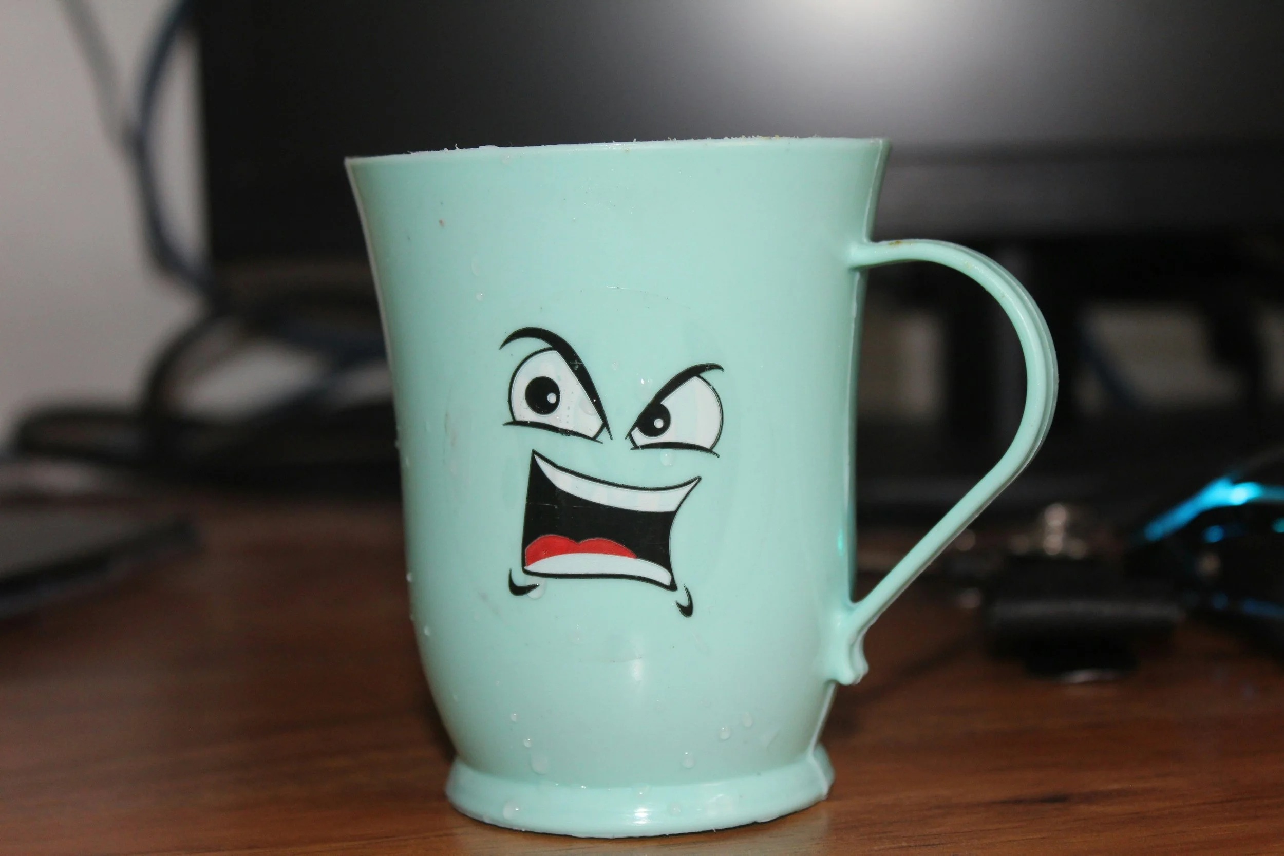 A light green mug with a cartoon face and angry expression, placed on a wooden surface.