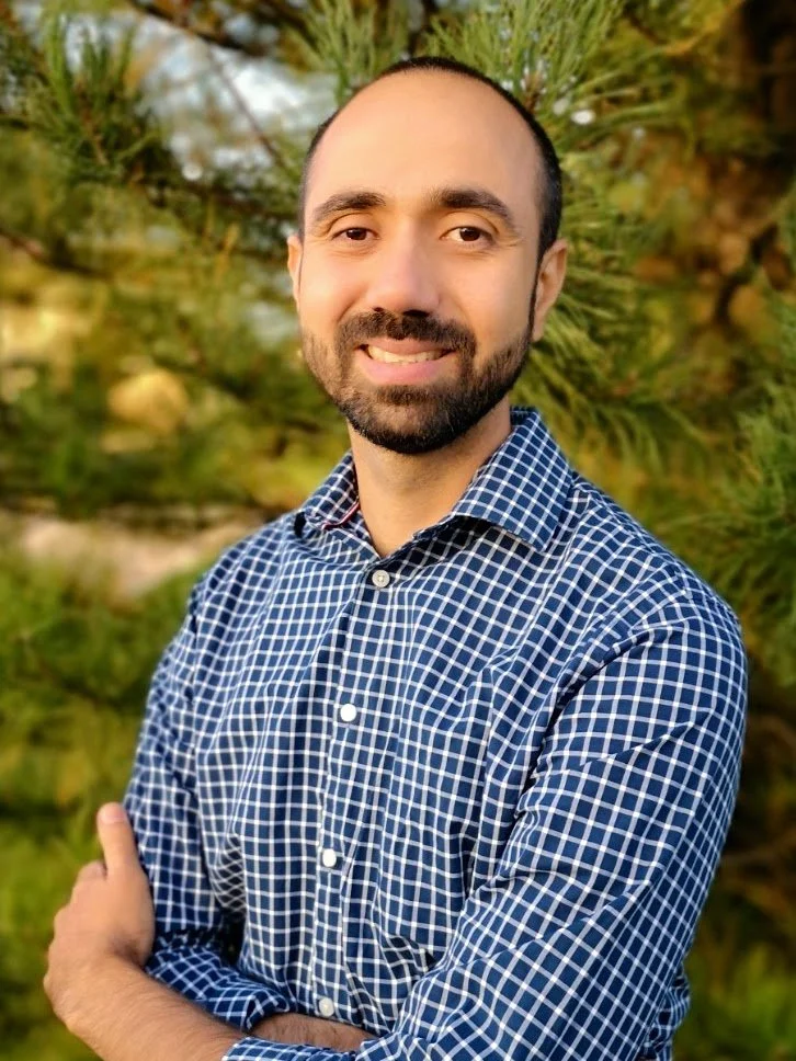 A man with a beard and short hair wearing a blue checkered shirt standing outdoors with green trees in the background.