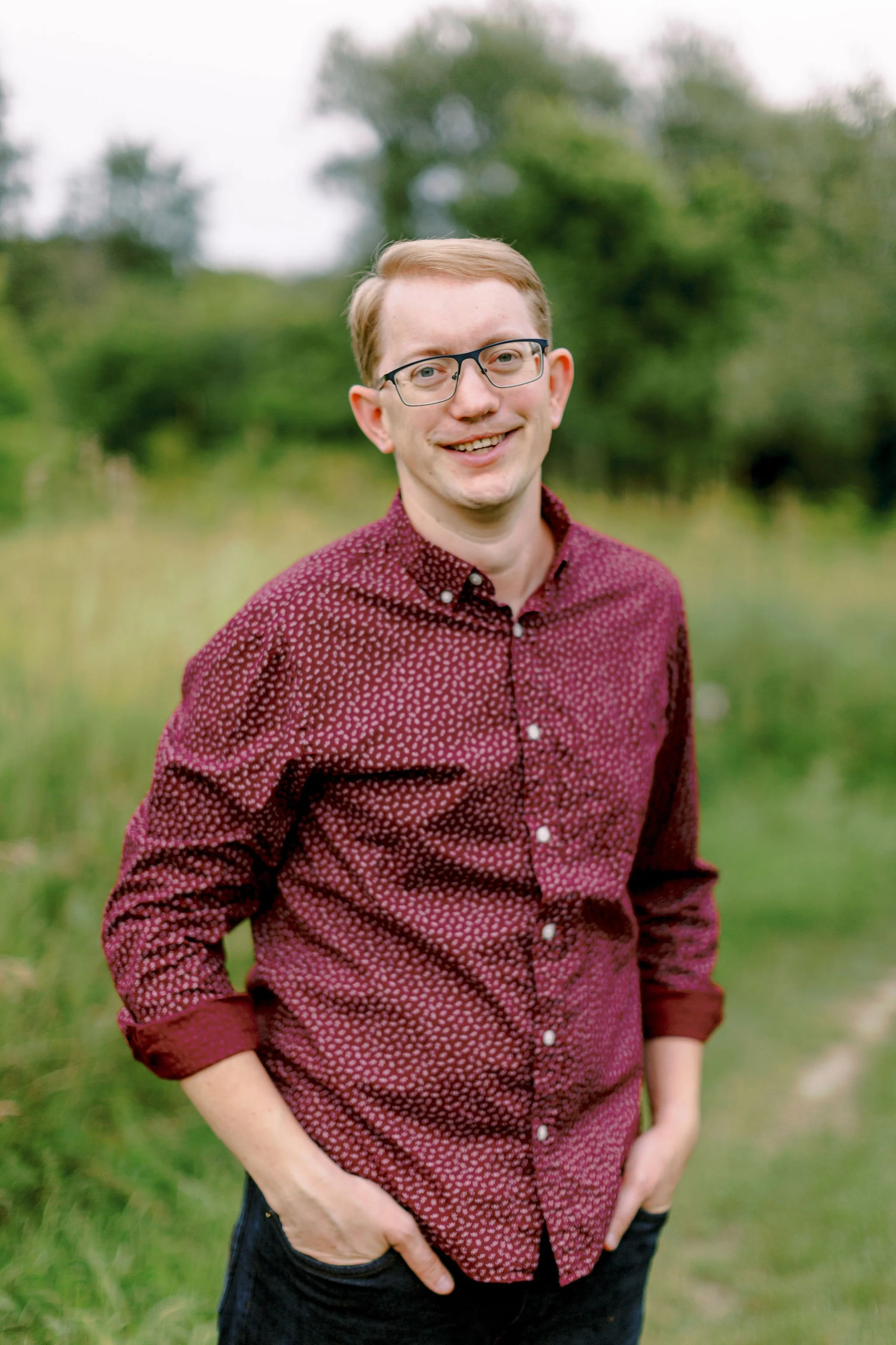 A young man with glasses, light hair, and a friendly smile, wearing a maroon patterned shirt, standing outdoors in a green field with trees in the background.