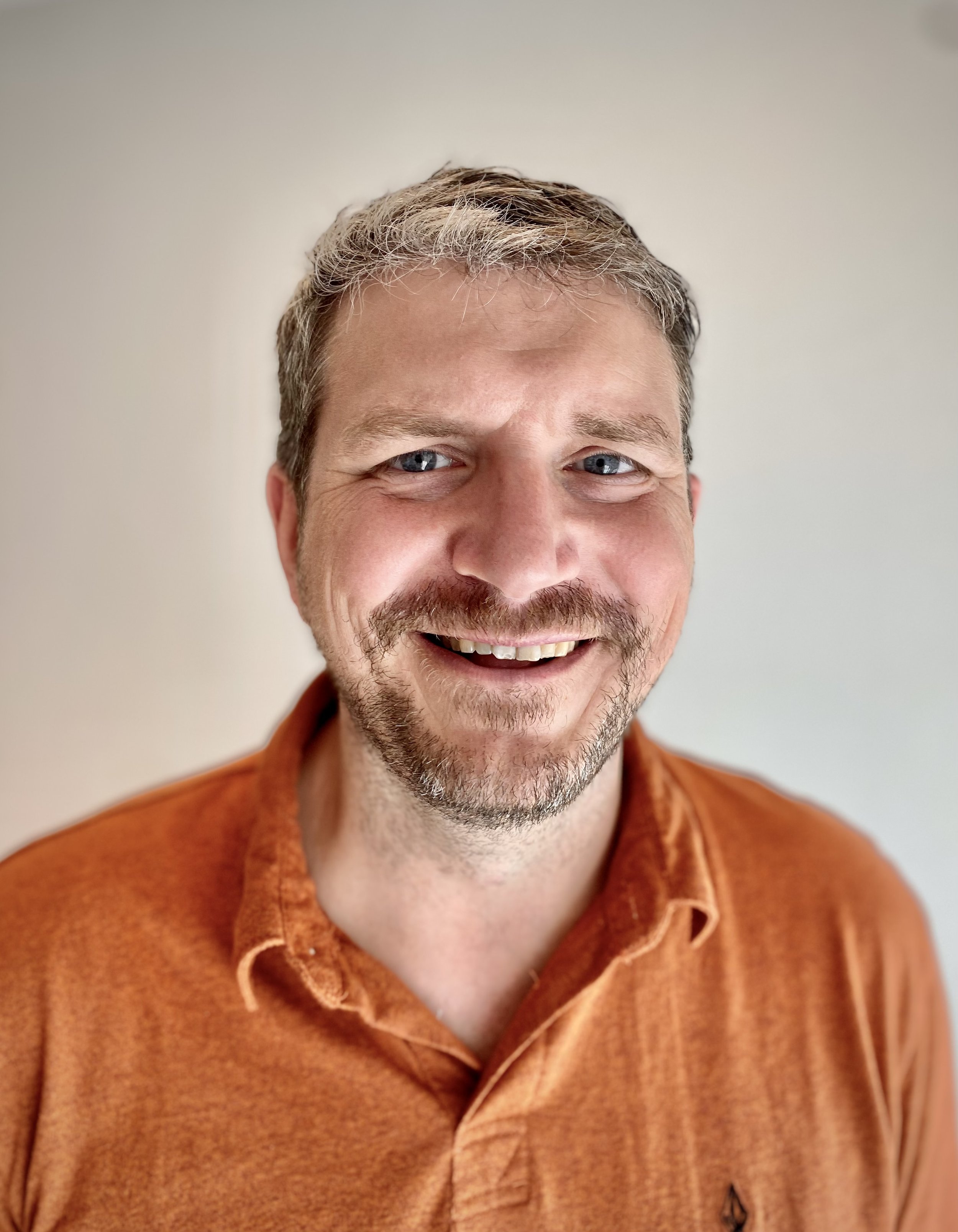 Close-up of smiling man with short, curly hair and blue eyes, wearing an orange shirt, against a plain light background.