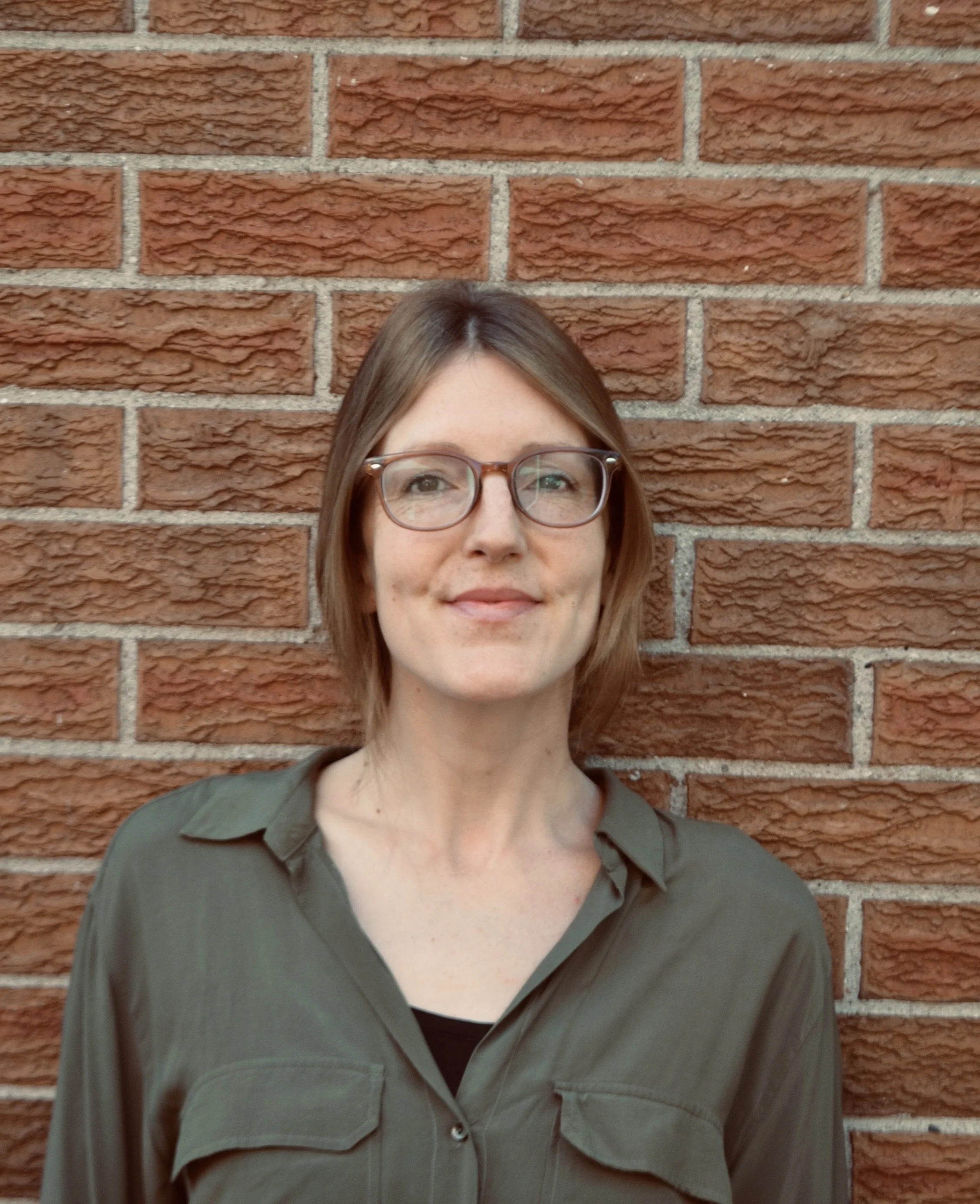 Close-up of a woman with glasses smiling, standing in front of a red brick wall.