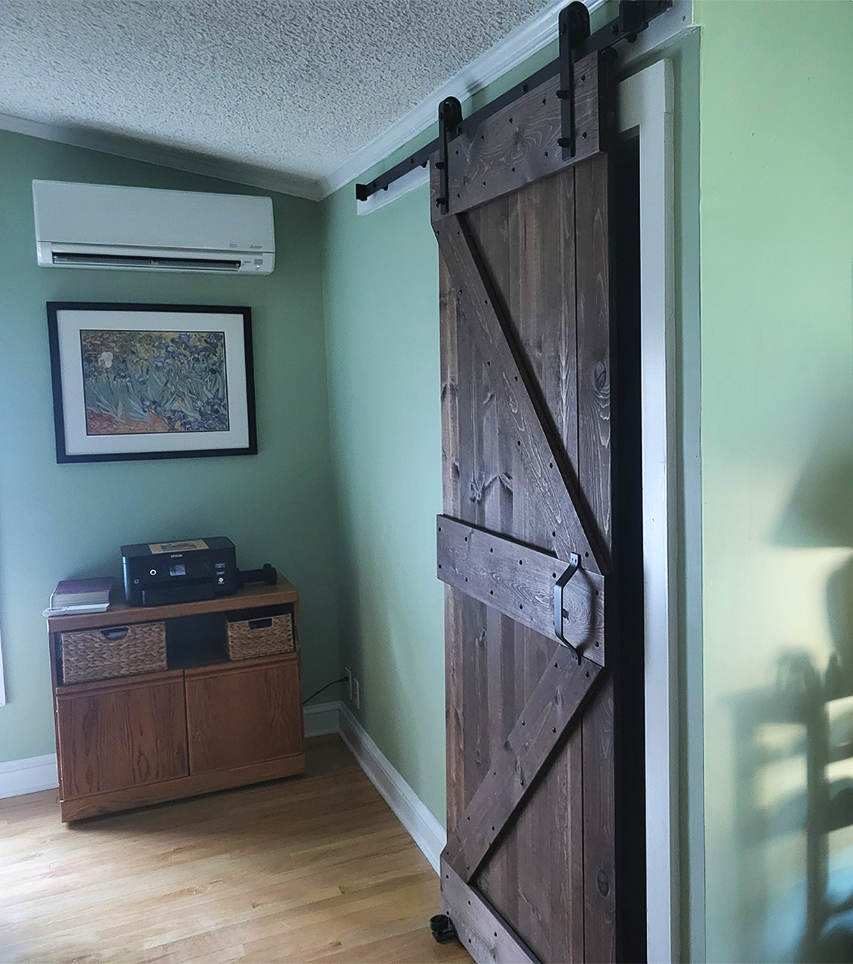 A wooden sliding barn door on a black track mounted on the wall inside a house. To the left, there's a small wooden cabinet with a printer on top. Above the cabinet, a framed artwork hangs on a pale green wall, and a white air conditioning unit is mounted near the ceiling.