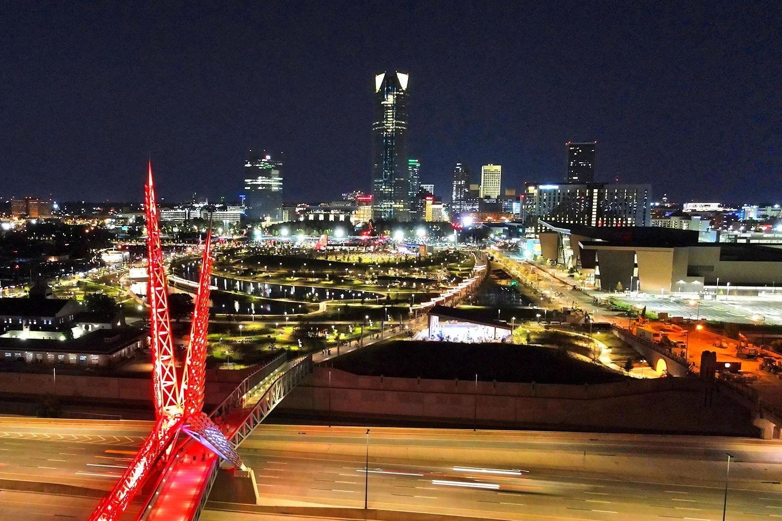 Nighttime city skyline with illuminated skyscrapers, a modern bridge with red lights, and a large arena in the city of Oklahoma City.