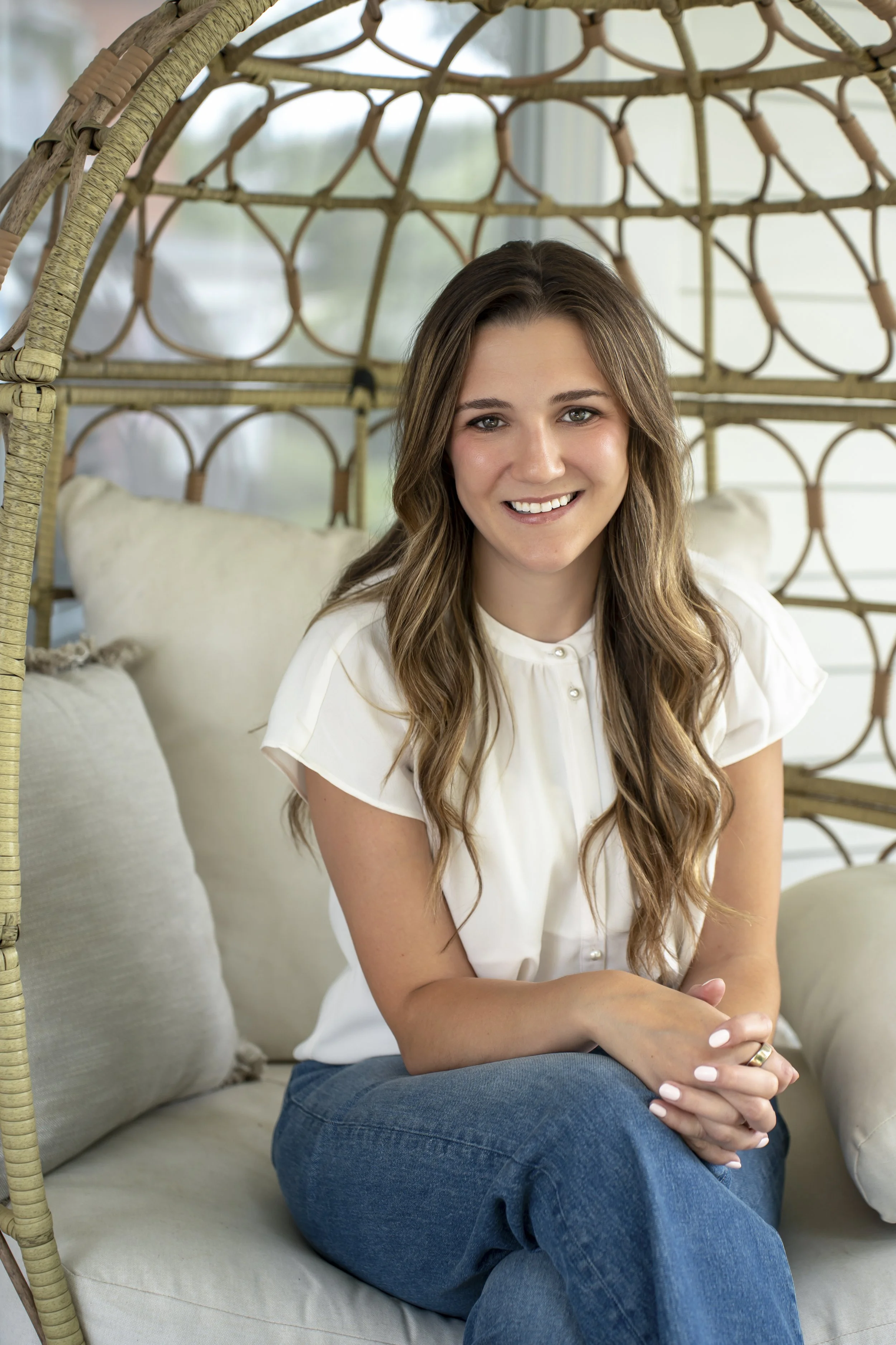 A young woman with long wavy brown hair sitting on a beige sofa, smiling, with a wicker canopy behind her and a window with white siding in the background.