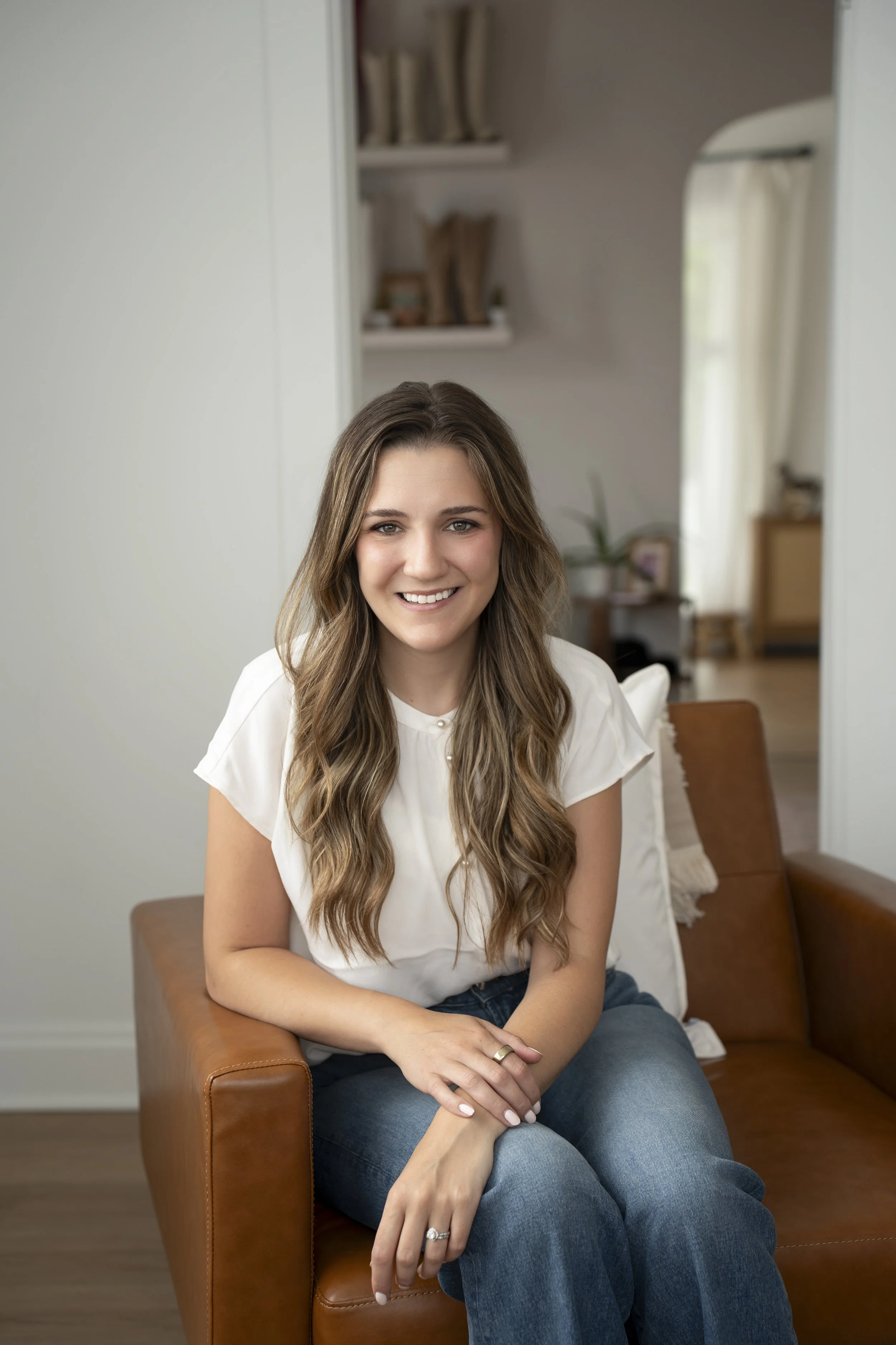 A woman with long wavy brown hair smiling, sitting on a brown leather armchair in a cozy room with white walls, a small table, bookshelf, and a mirror in the background.