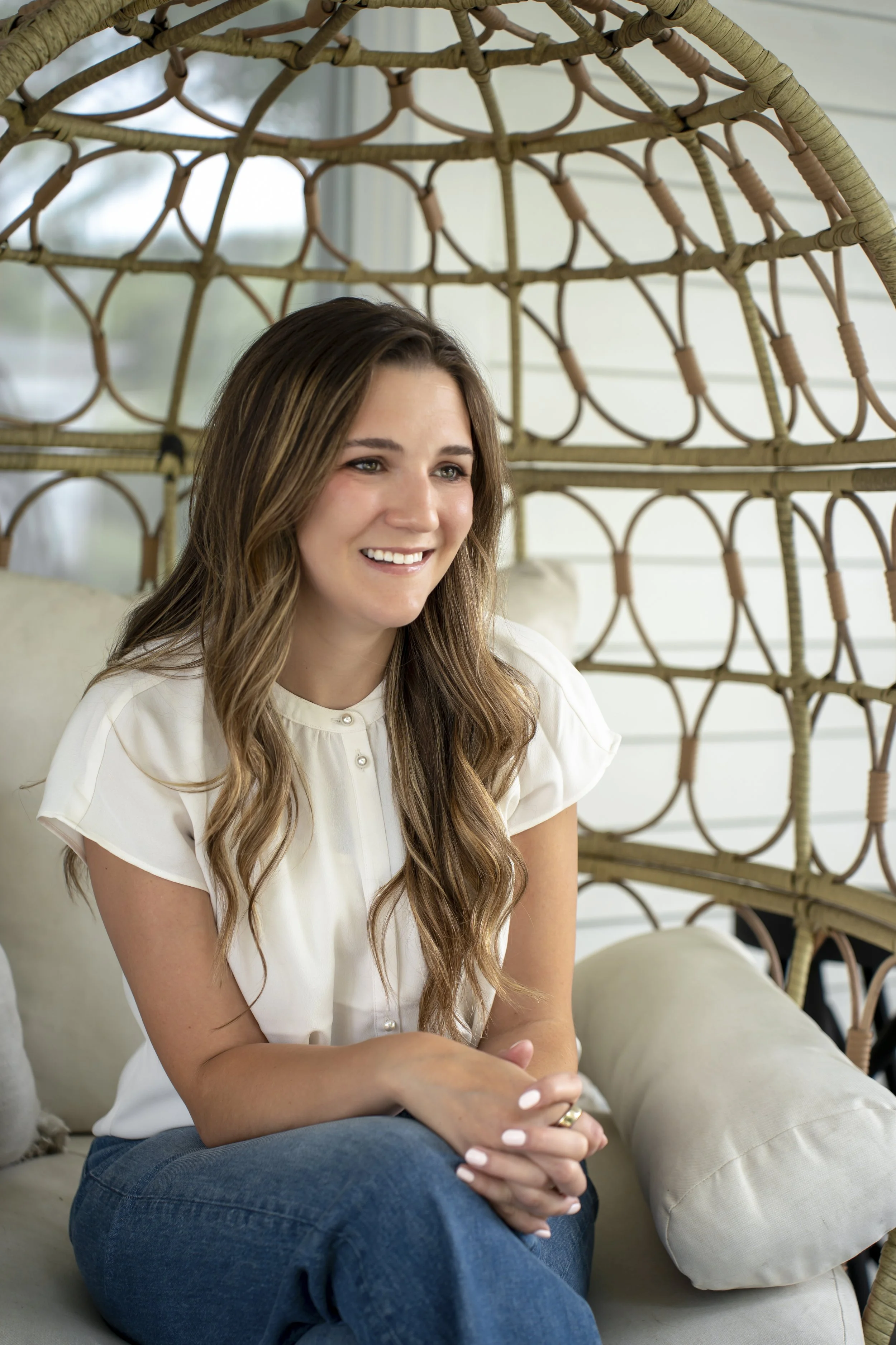 A young woman with long wavy brown hair, wearing a white blouse with pearl buttons and blue jeans, sitting on a cream-colored couch in a bright room with white walls and large windows. She is smiling and looking content.