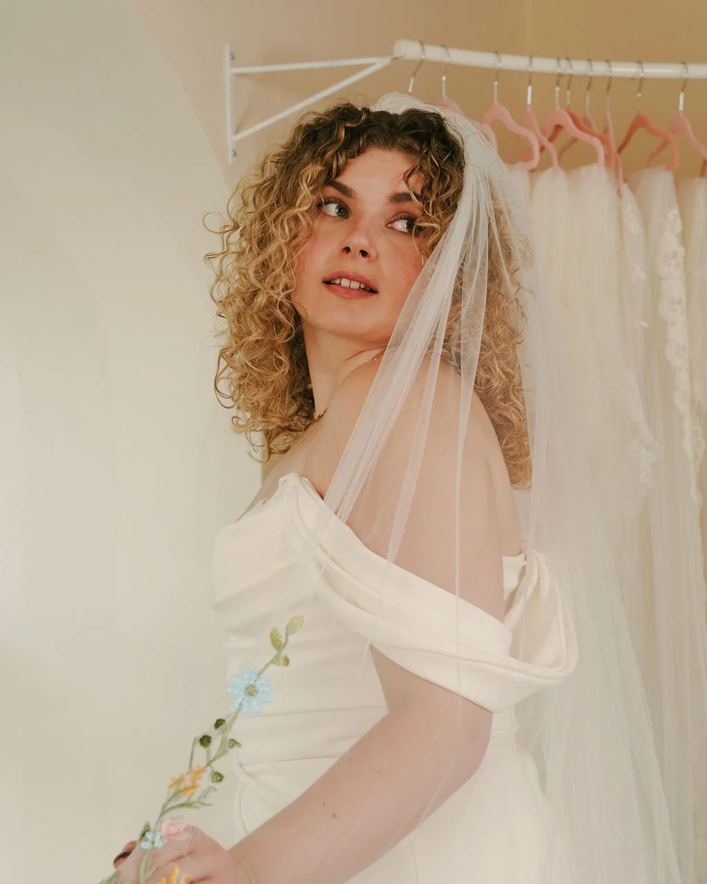 A woman with curly blonde hair wearing a white wedding dress with floral embroidery, looking over her shoulder with a veil on her head, standing in front of a clothing rack with bridal dresses.