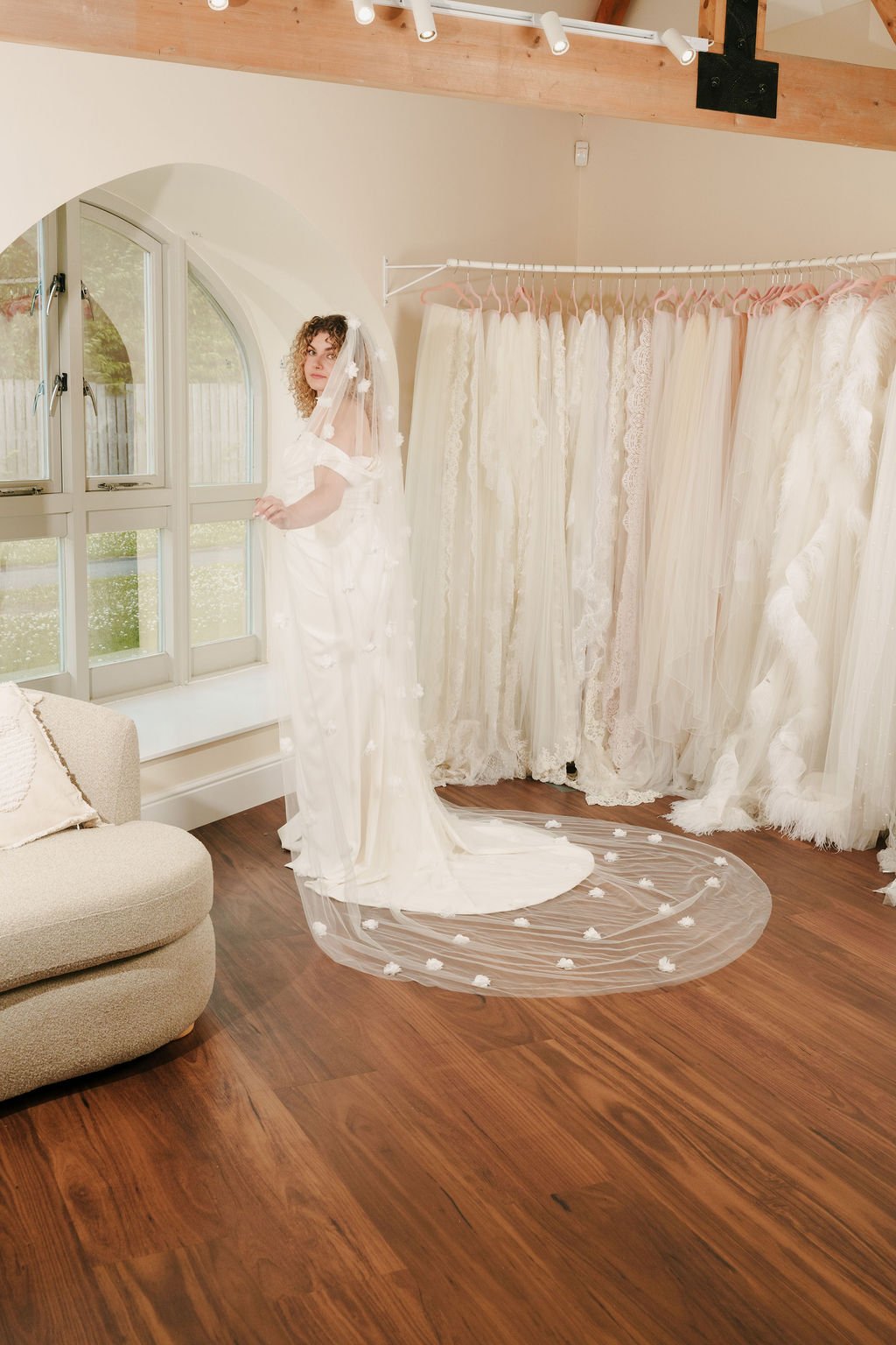 A woman browsing wedding dresses inside a boutique, standing in front of a large window with a view of greenery, wearing a white wedding gown and veil, with lace dresses hanging on the wall behind her.