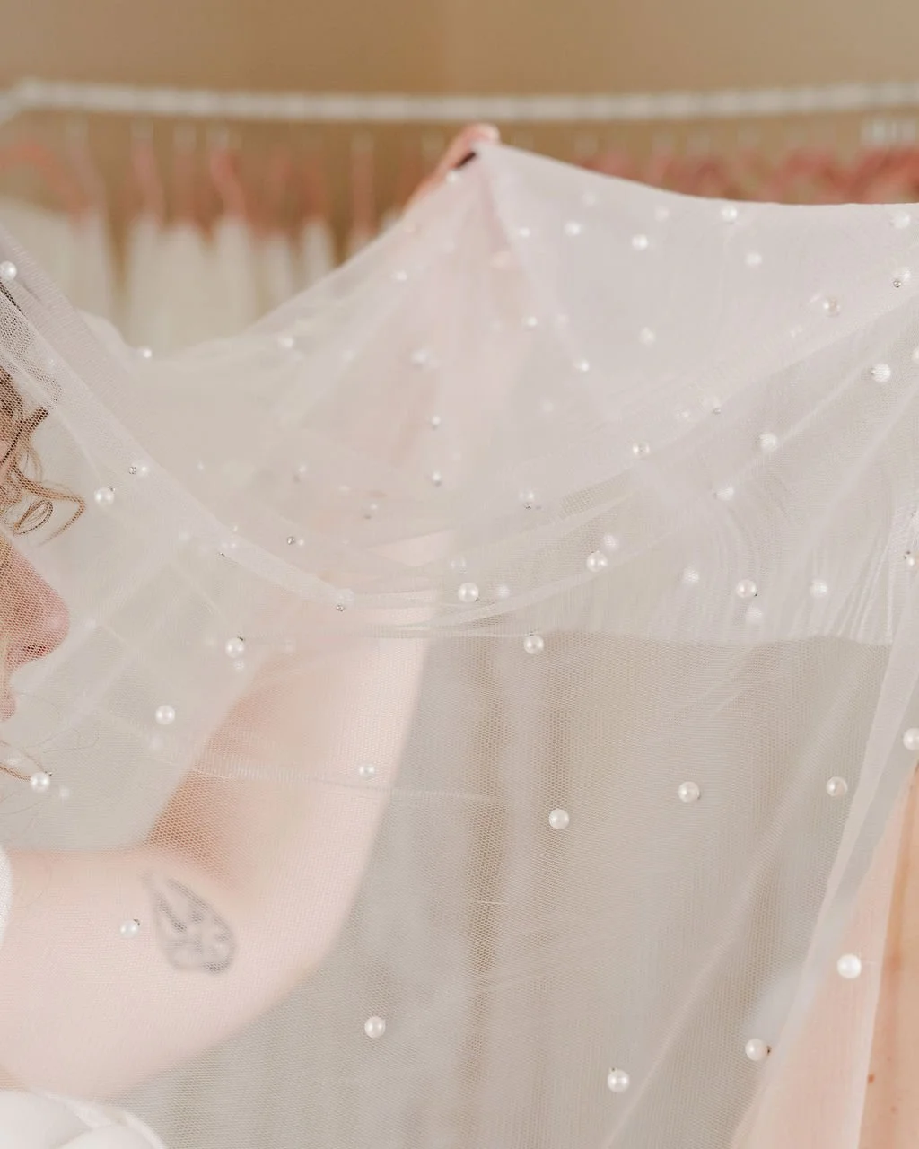 Close-up of a person holding a delicate, sheer veil with pearl embellishments in a clothing store or boutique, with clothing racks in the background.