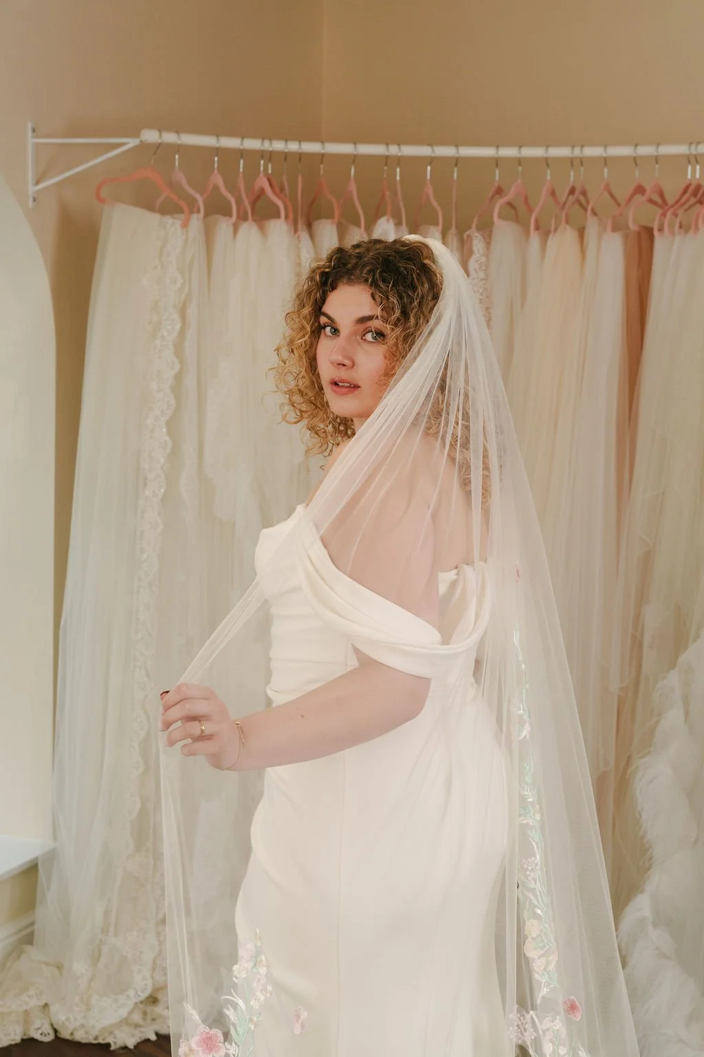 A woman trying on a wedding dress with sheer veil in a bridal shop, with other wedding gowns hanging in the background.