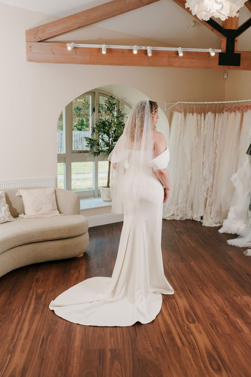 A bride in a white wedding dress with a veil stands in a bright, elegant room with hardwood floors. Behind her, a window and a potted plant are visible, along with a curtain display of various white dresses.