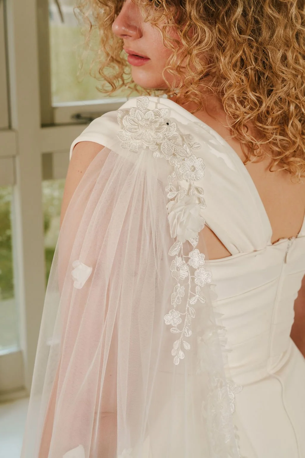 Close-up of a woman with curly blonde hair wearing a white dress with floral lace appliqué and a sheer cape with floral embroidery, standing near a window.