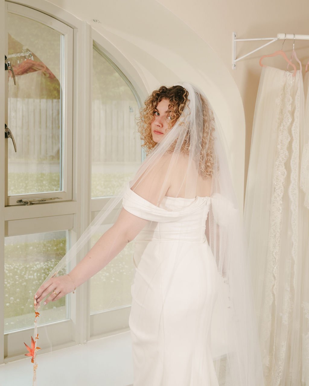 A woman with curly hair wearing a wedding dress and veil, standing near a window inside a room with hanging wedding dresses.