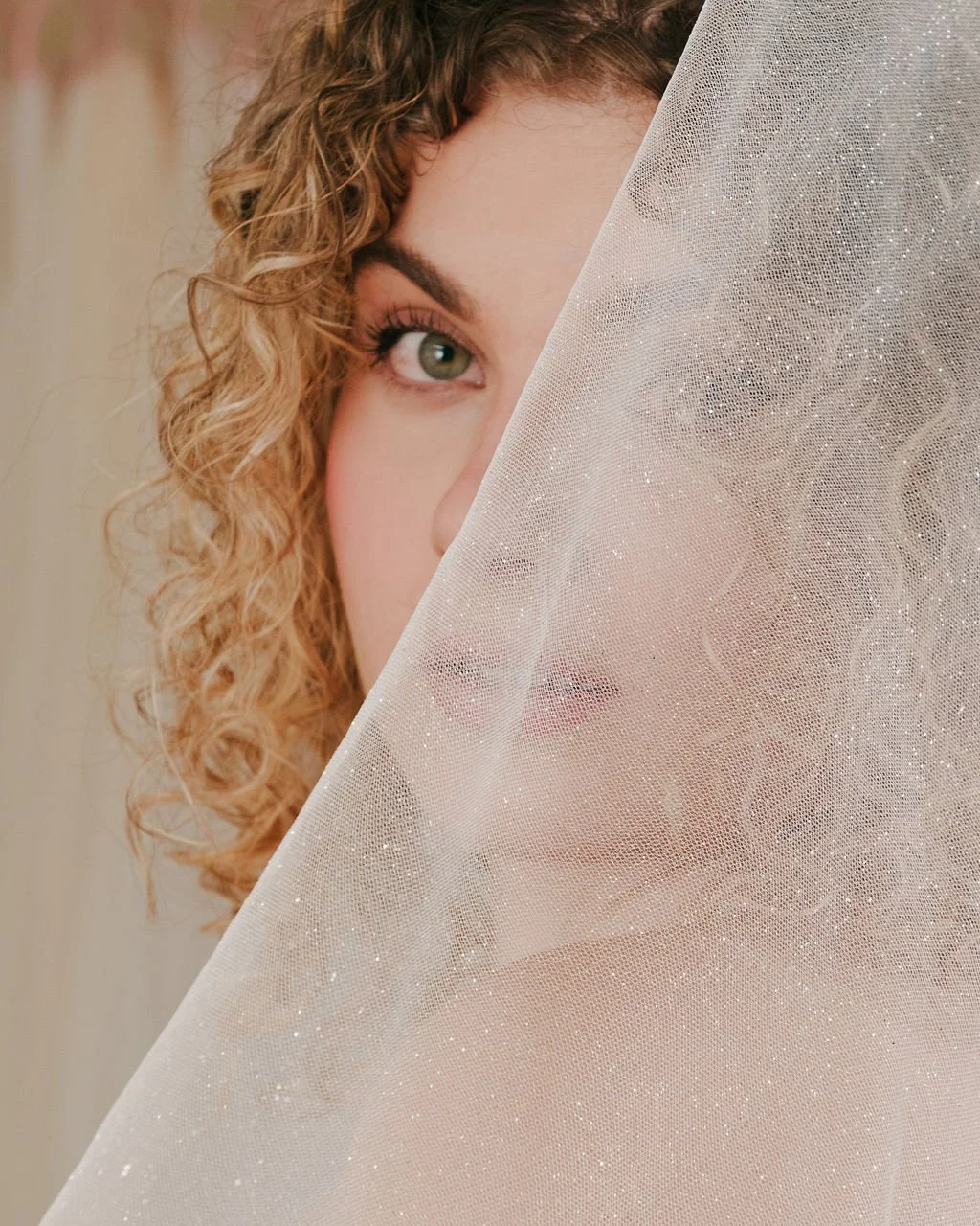 A woman with curly red hair and green eyes peeks through a sheer, glittery veil covering part of her face.