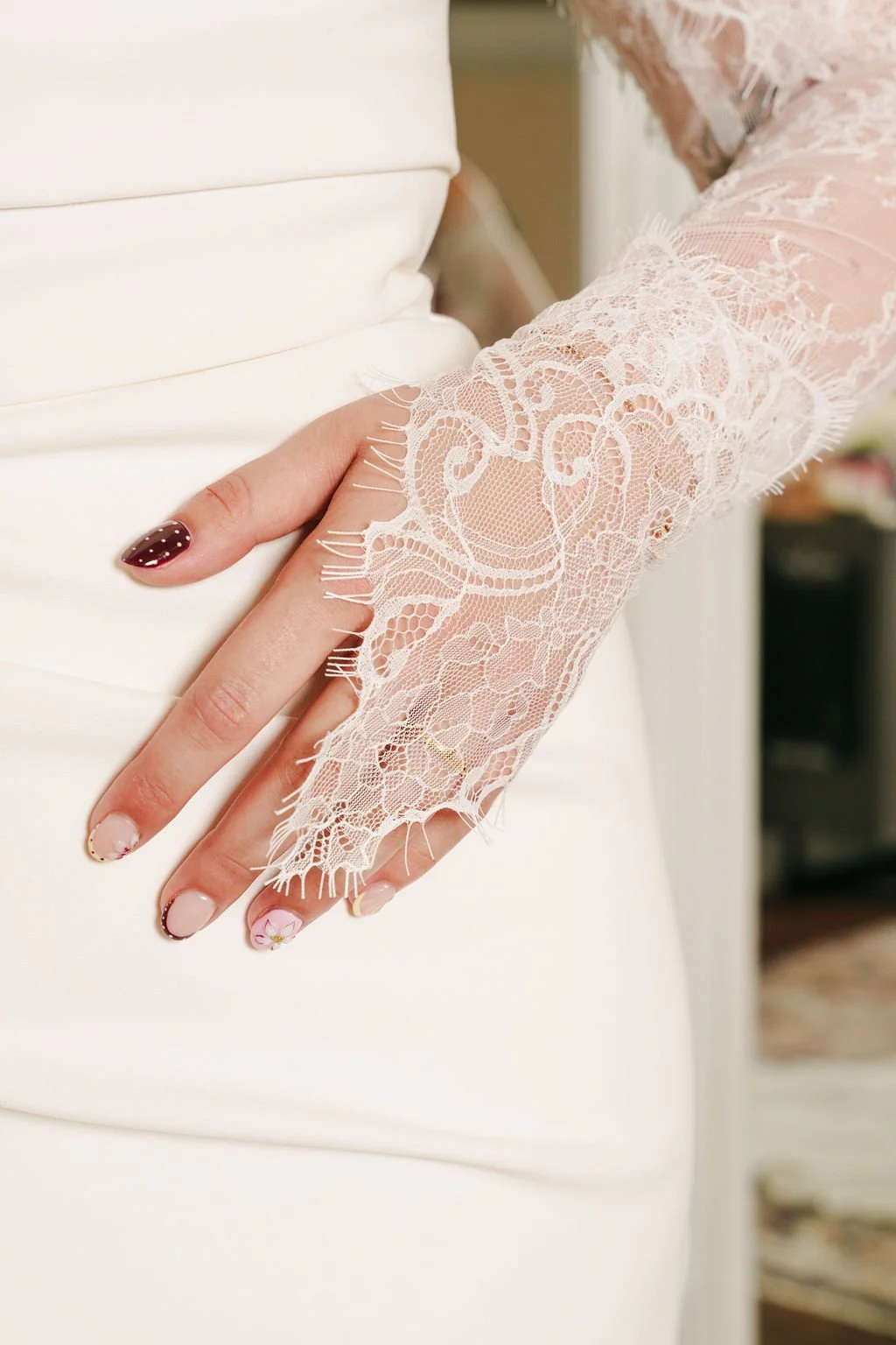 Close-up of a bride's hand with manicured nails, resting on her white wedding dress. The wedding dress features intricate lace with floral patterns and fringe details on the sleeve.