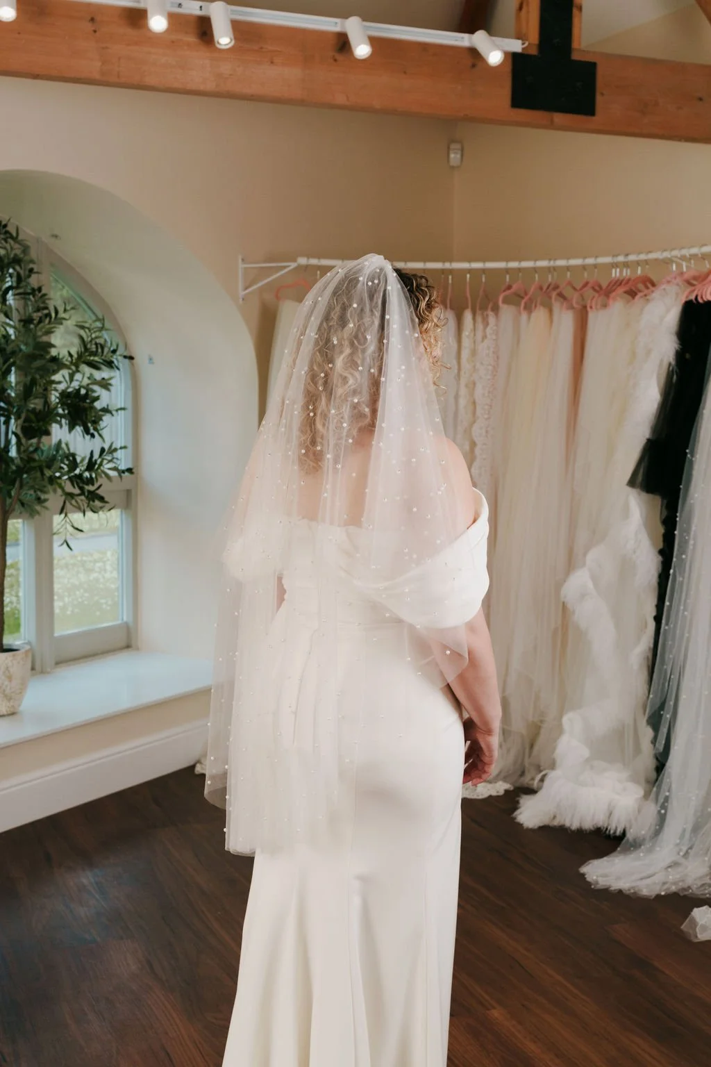 Woman trying on a wedding dress with veil in a bridal shop surrounded by other wedding gowns.