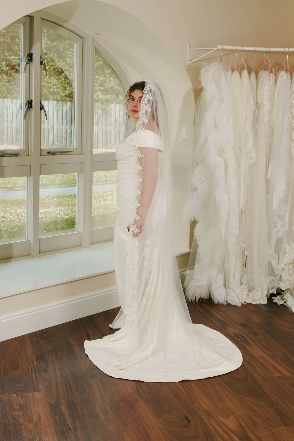 A woman in a white wedding dress and veil stands inside a room next to a large window with a view of greenery outside. There are several white wedding dresses hanging on a rack in the background.