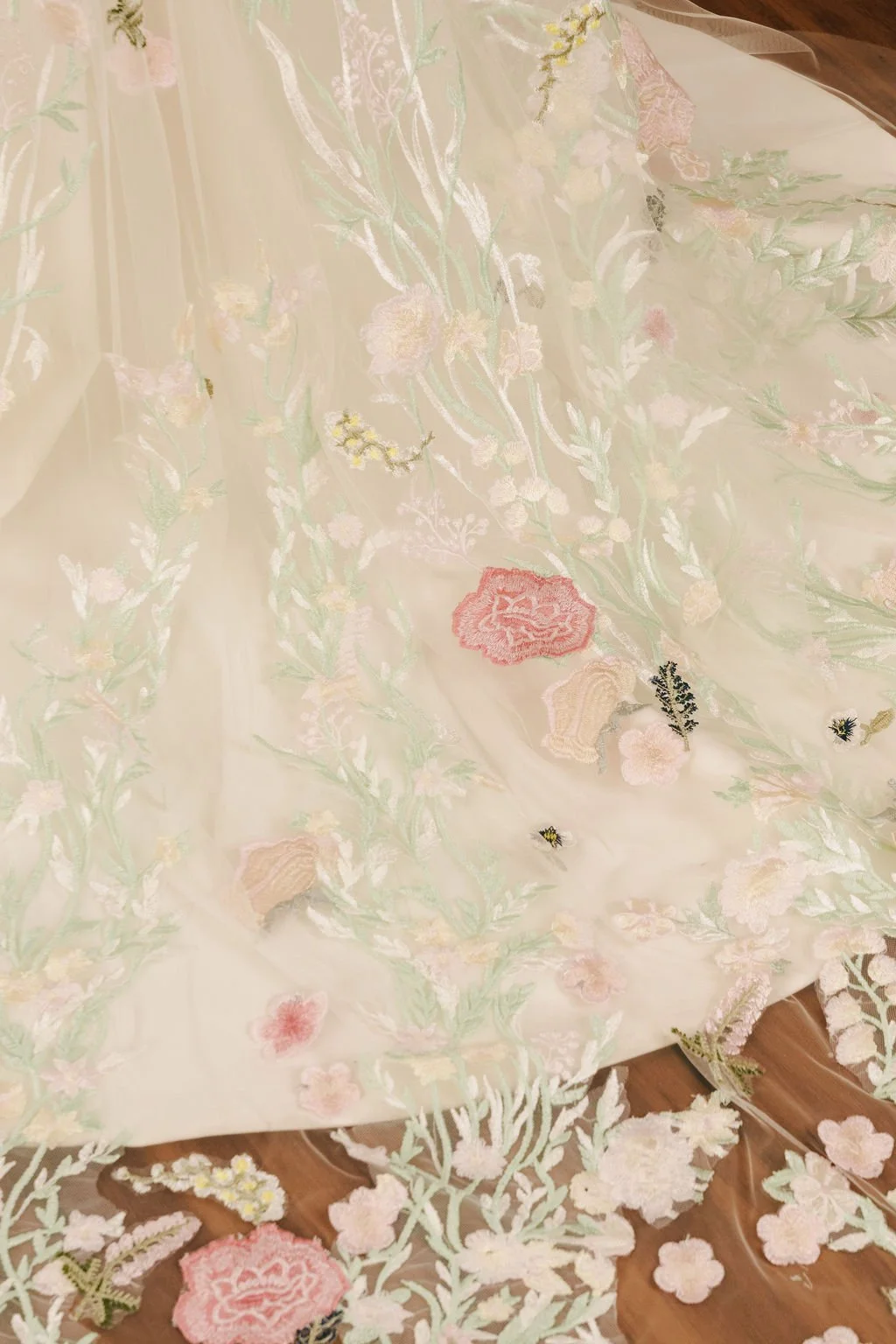 Close-up view of a light-colored fabric with intricate floral embroidery in pastel shades, including pink, yellow, green, and cream. Some embroidered flowers and leaves are on a wooden surface.
