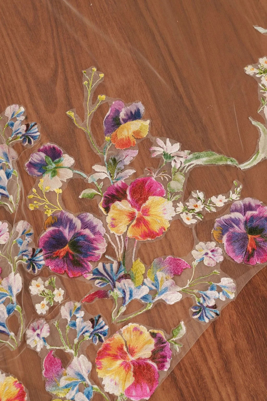 Colorful embroidered floral design on sheer fabric with various flowers in pink, purple, yellow, white, and blue, on a wooden surface.
