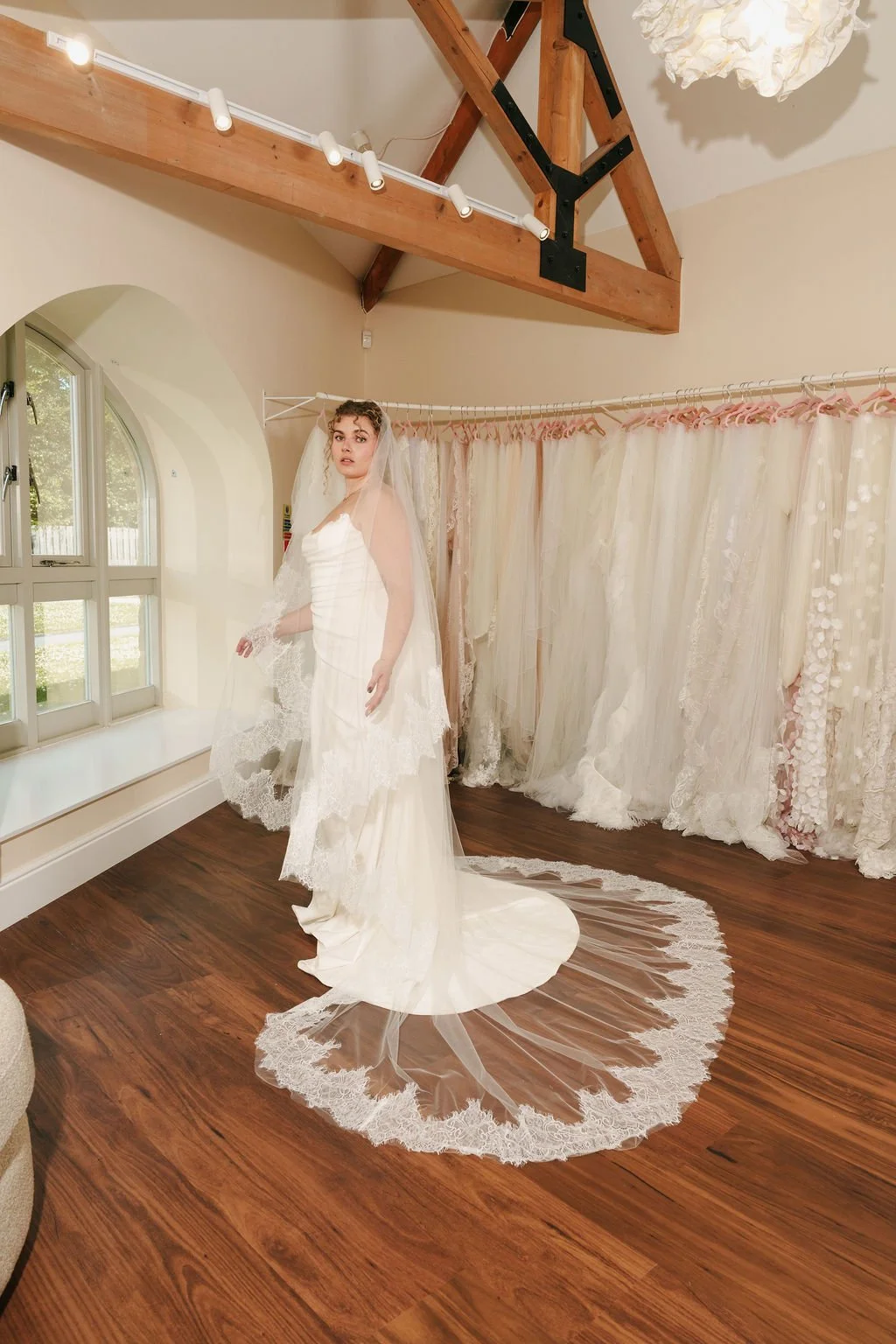 A woman in a wedding dress with a veil stands on a wooden floor in a bridal boutique, with a curtain of various lace and fabric wedding dresses in the background and a window to the left.