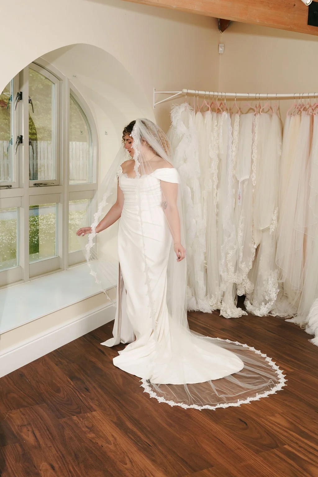 Bride trying on a wedding dress in a bridal shop with lace dresses hanging on a rack behind her.