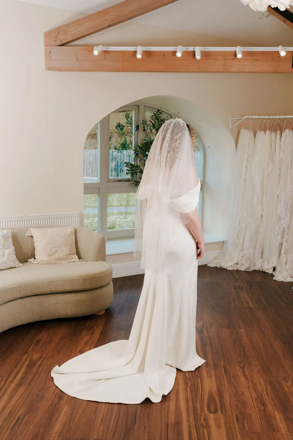 A woman dressed in a white wedding gown and veil standing in a bright room with wooden floors, a beige couch, a window, and a curtain of lace.
