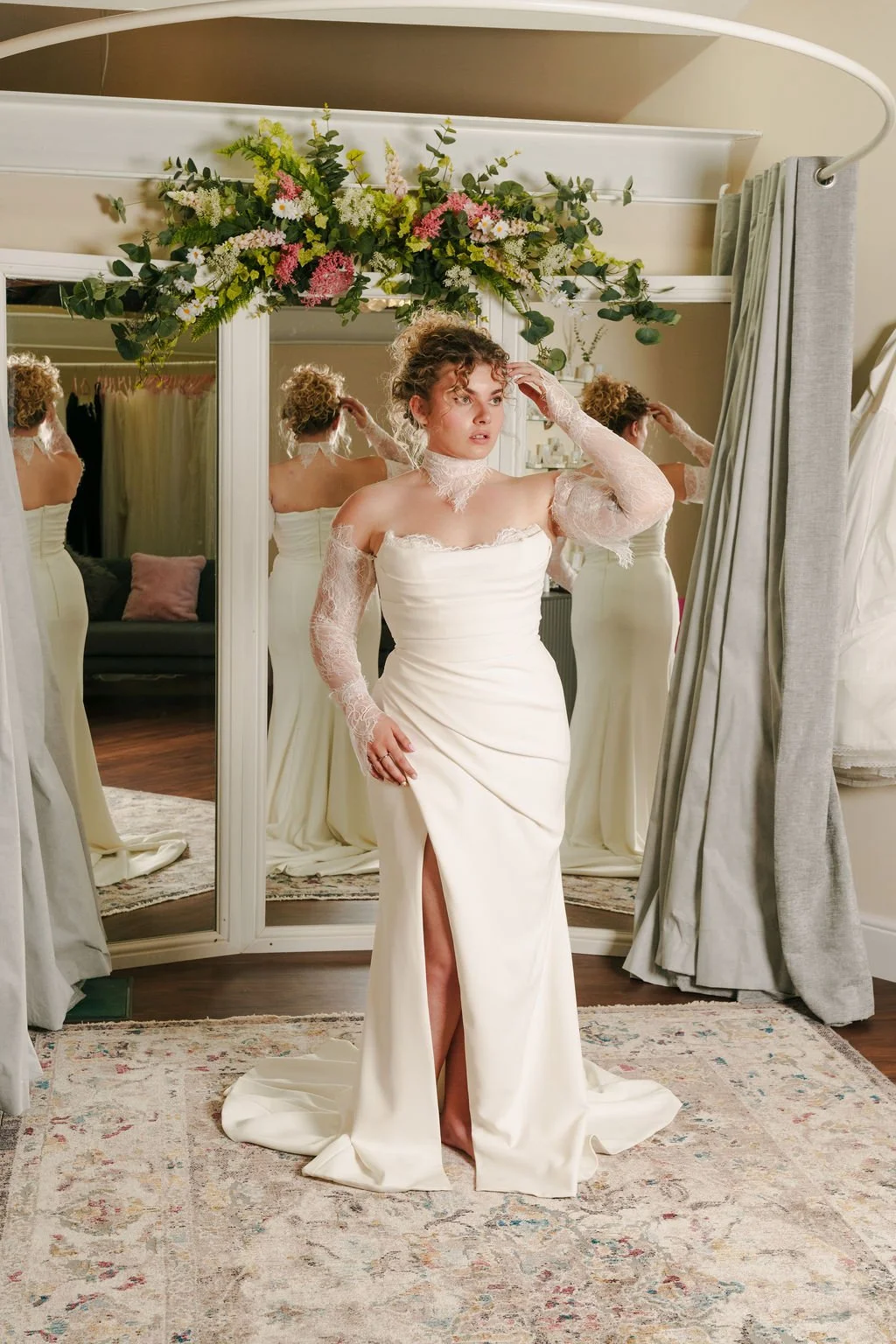 A woman in a white wedding dress with lace sleeves and a slit, adjusting her hair in front of a mirror adorned with a floral arrangement.