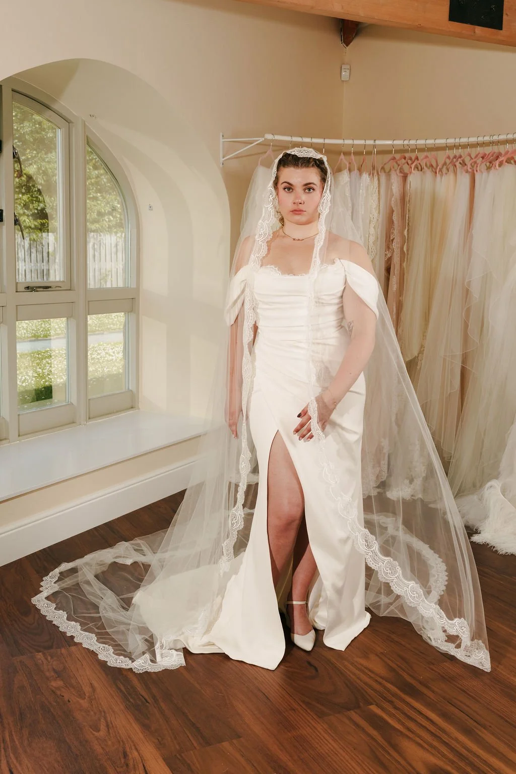 A bride trying on a white wedding gown in a bridal boutique, with window and fabric curtains in the background.