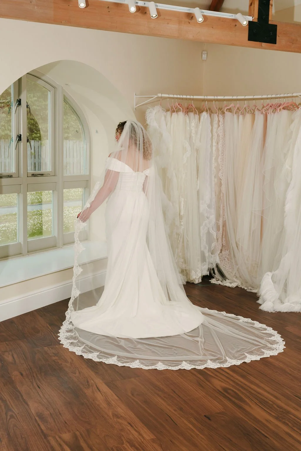 A bride in a white wedding dress with a veil stands in a bridal boutique, looking at herself in a mirror. She is surrounded by a rack of hanging wedding gowns with lace and tulle details. The room has wooden floors and large windows with a view of gr