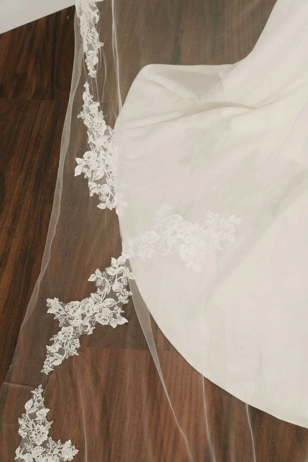 Close-up of a wedding veil with floral lace embroidery, lying on a wooden surface next to a white wedding dress.