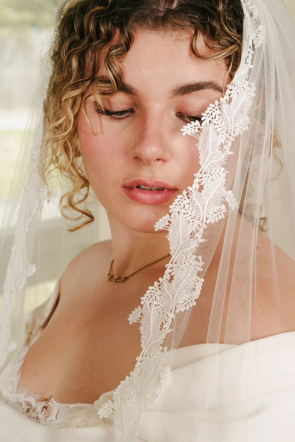 A close-up of a woman with curly hair wearing a white wedding dress and veil, standing near a window with soft natural light.