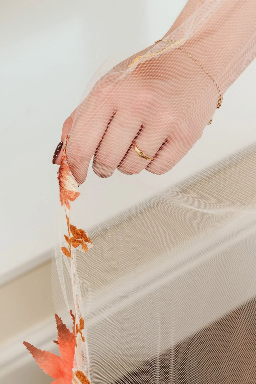 Close-up of a hand with several rings, holding a sheer fabric with orange and red embroidery and a leaf pattern, against a white background.