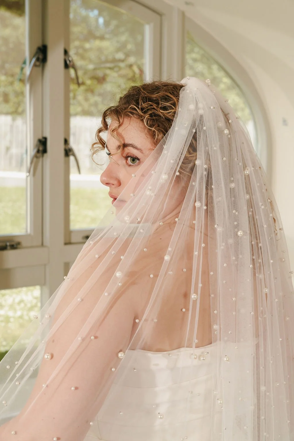A woman with curly hair wearing a wedding dress and a veil decorated with pearls, looking over her shoulder near a window.