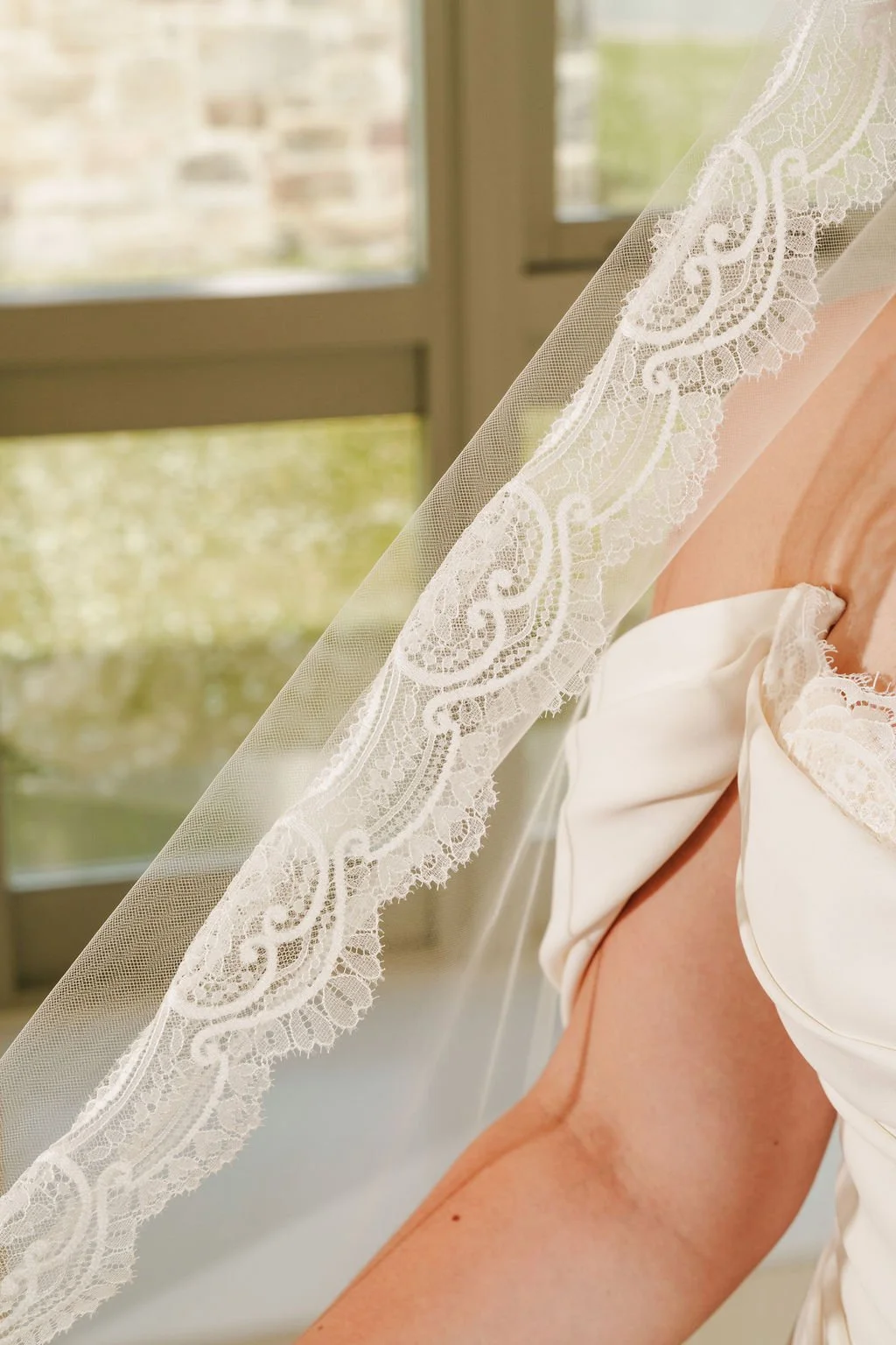 Close-up of a wedding dress with lace details on a woman sitting near a window with natural light.