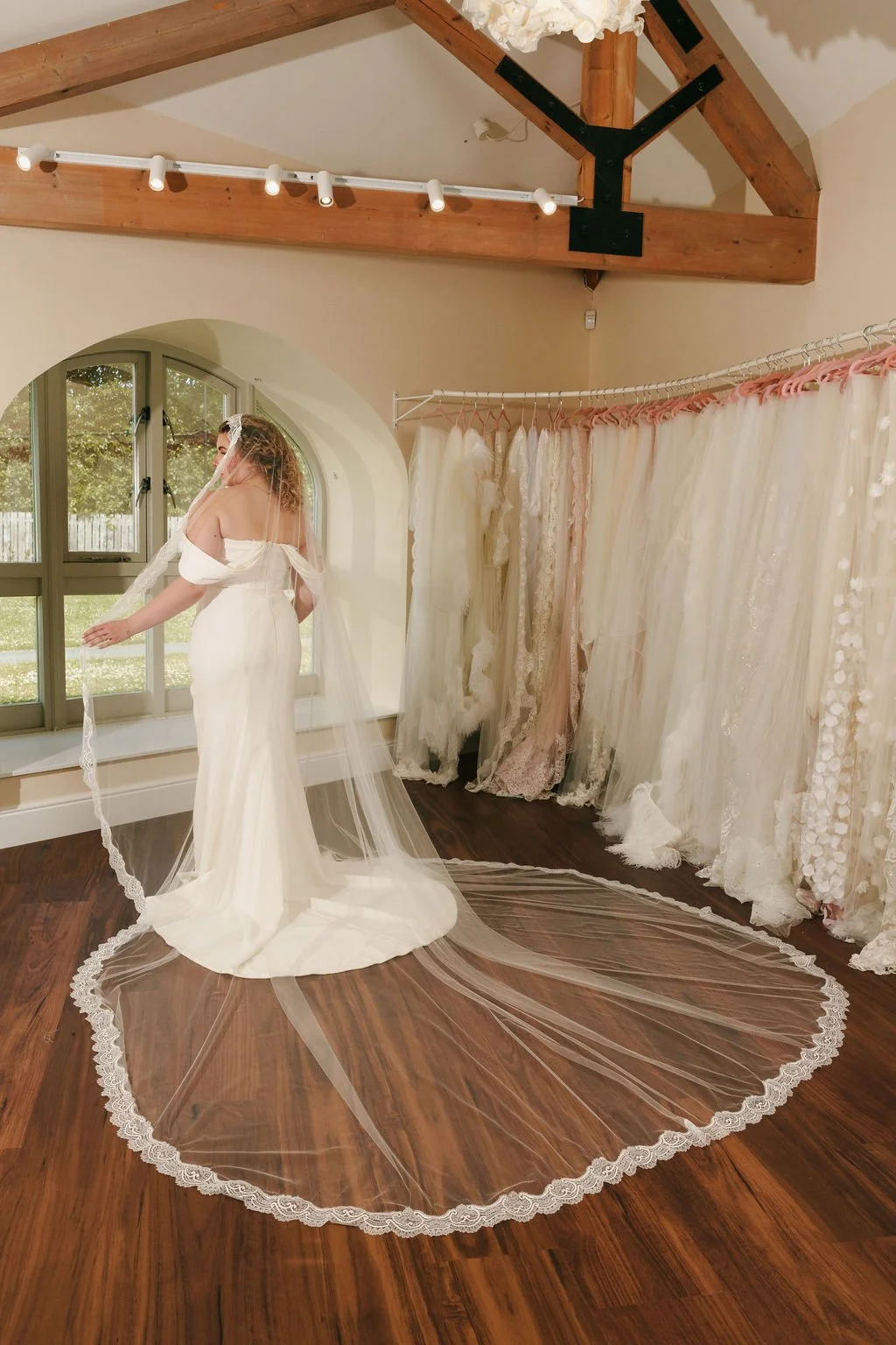 A woman in a white wedding dress trying on a veil in a bridal boutique with wedding gowns hanging on racks.