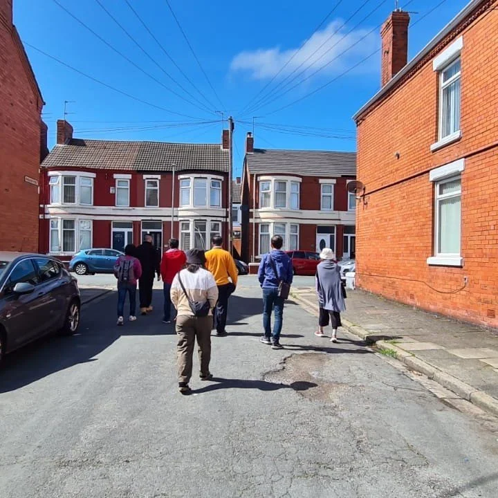 A group of people walking down a residential street with brick houses on both sides, parked cars, and wires overhead on a sunny day.