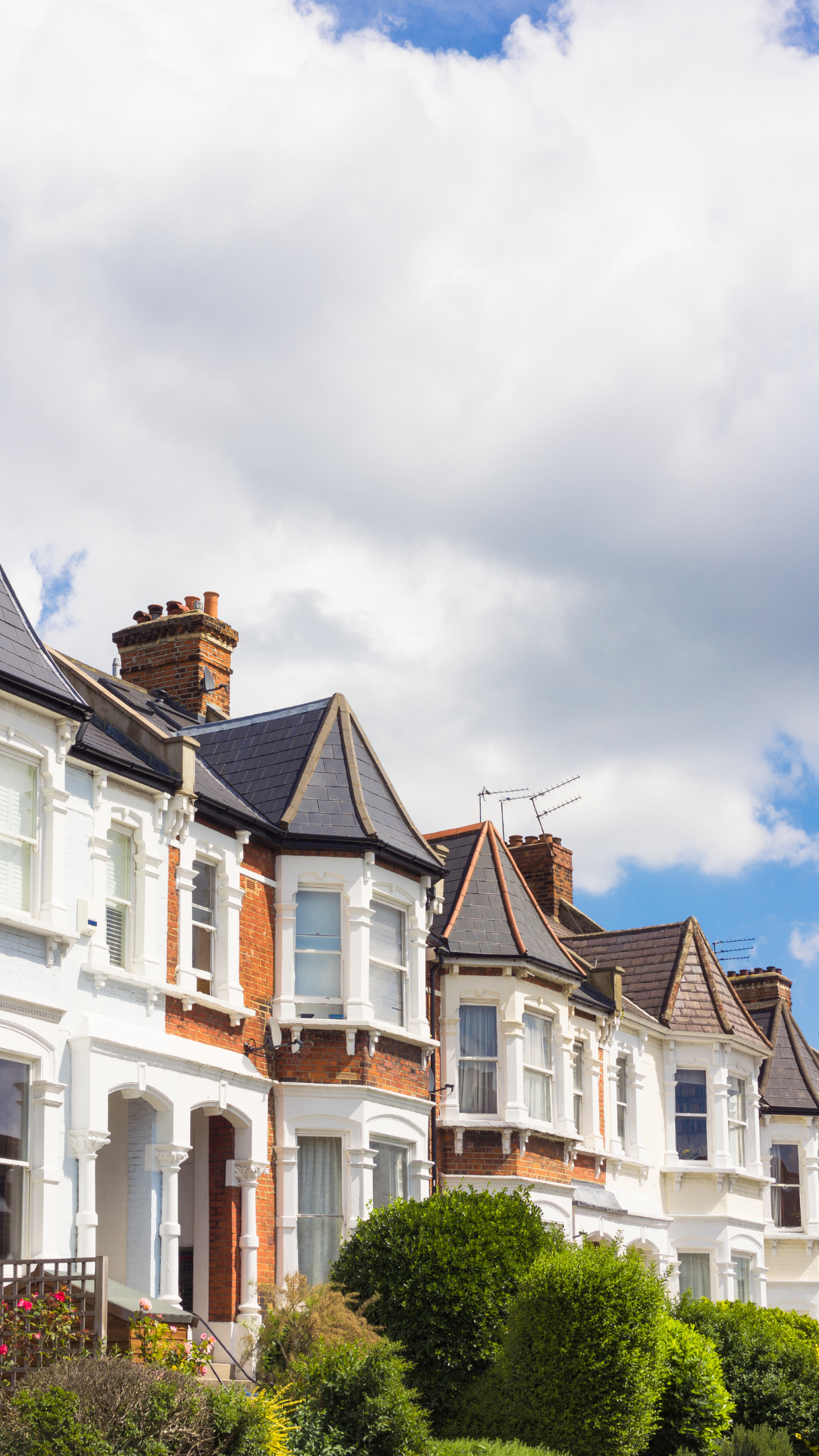 Row of Victorian-style houses with bay windows, brick and white exterior, surrounded by trees, under a partly cloudy sky.