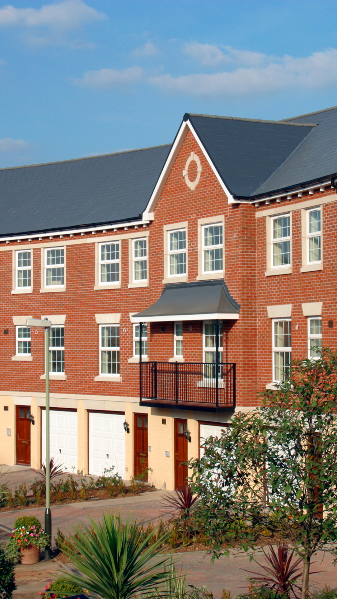 A red brick multi-story residential building with white window frames, a small balcony with black railing, and a gabled roof with a decorative round emblem. There is landscaping with plants and trees in front of the building and a streetlamp nearby.