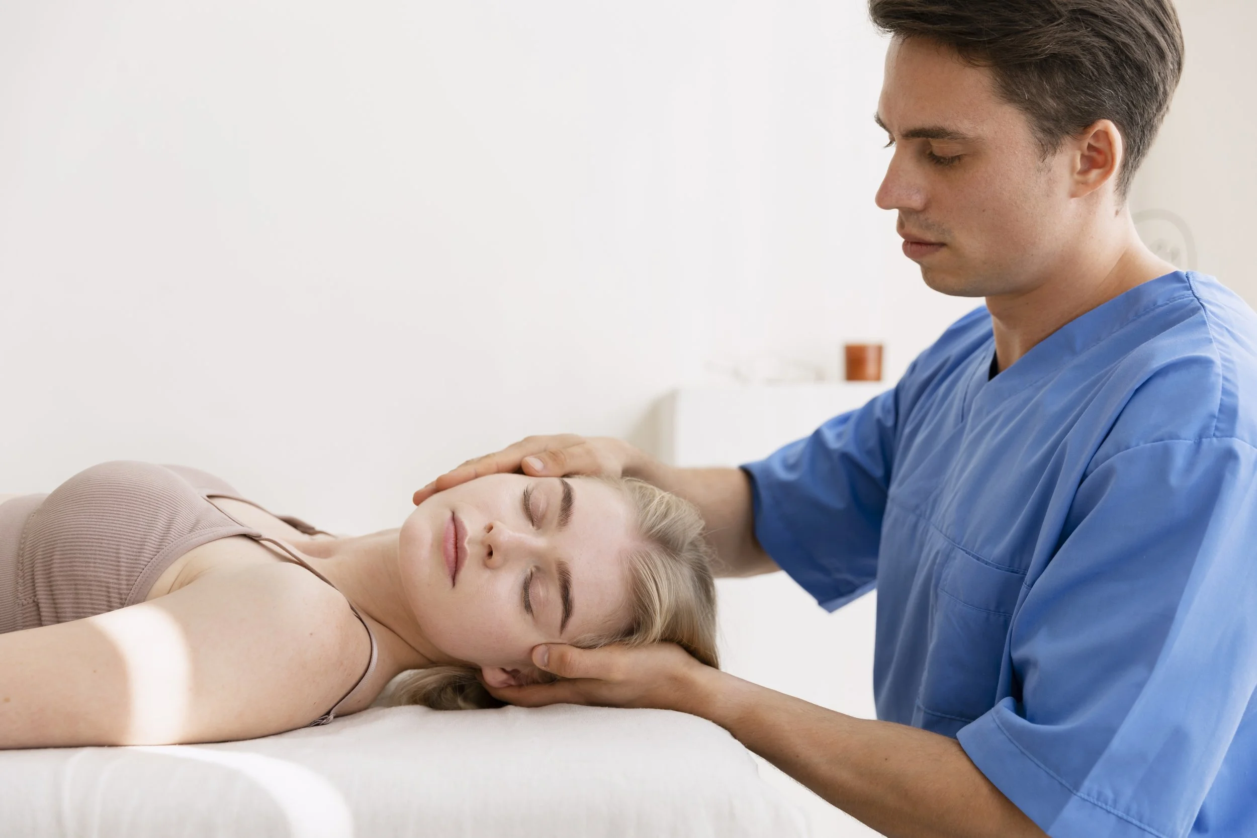 A healthcare professional in blue scrubs providing chiropractic care to a woman lying face-up on a white treatment table with her eyes closed, in a medical setting.