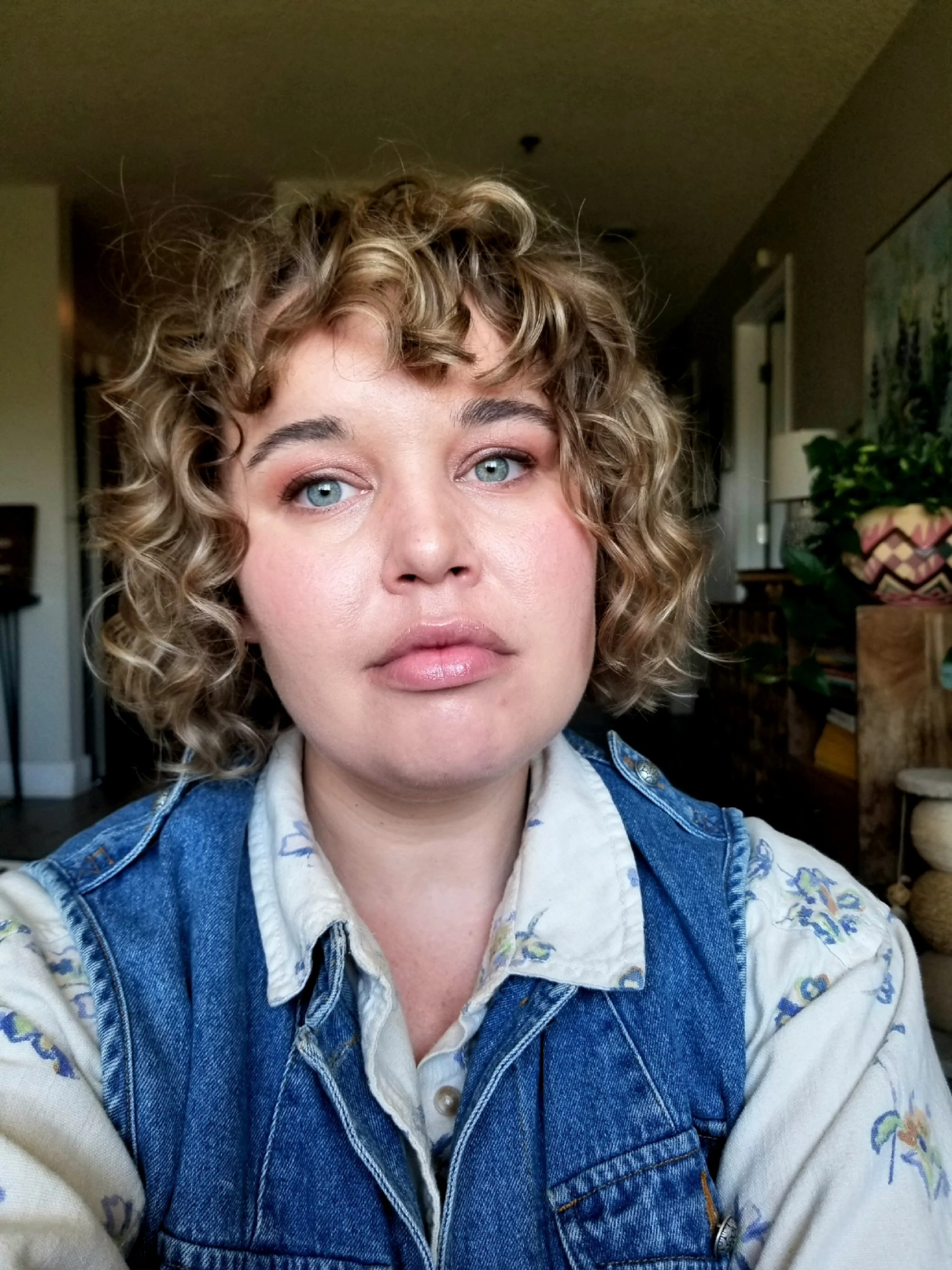 Close-up of a woman with blonde curly hair, wearing a denim vest over a floral shirt, in a room with plants and artwork in the background.