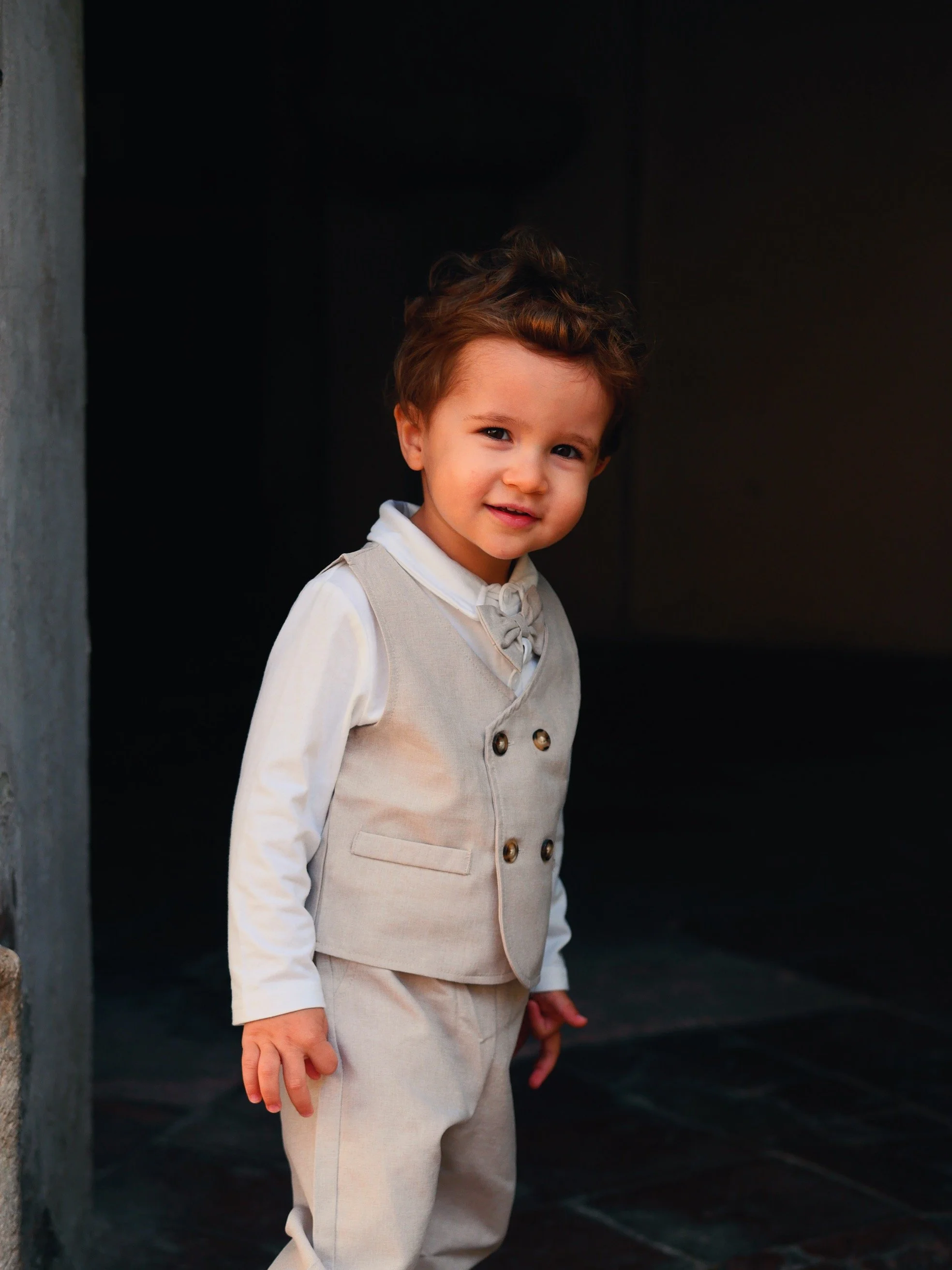Jeune garçon avec cheveux bruns bouclés, souriant, portant un gilet beige et une chemise blanche, debout près d'un mur sombre.