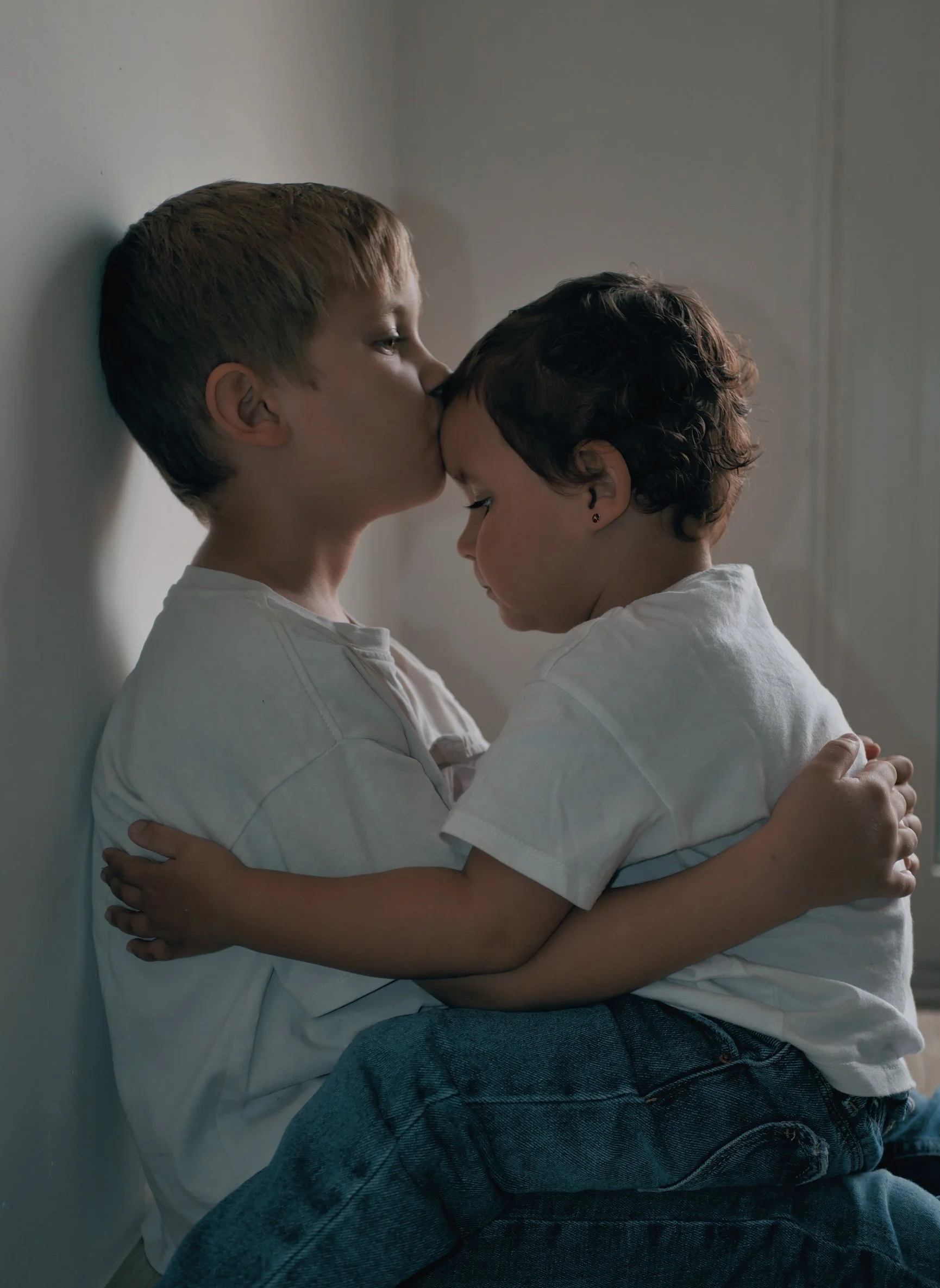 Deux enfants, un garçon plus âgé et un garçon plus jeune, se donnent un baiser sur le front dans une pièce à l'intérieur.