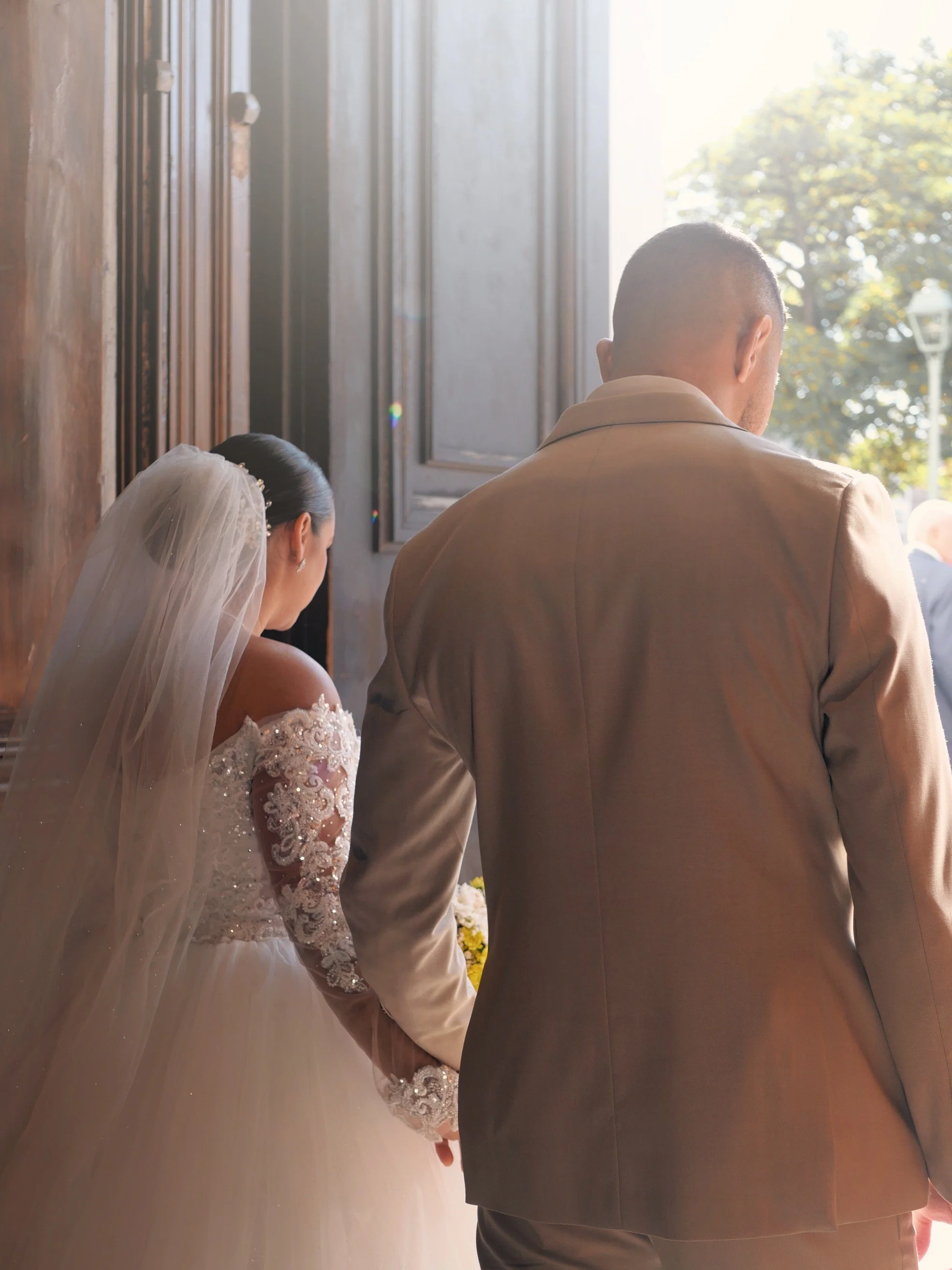 Un couple de mariés se tenant par la main, le dos, dans une pièce lumineuse avec des fenêtres en arrière-plan, échangeant leurs vœux lors de leur mariage.
