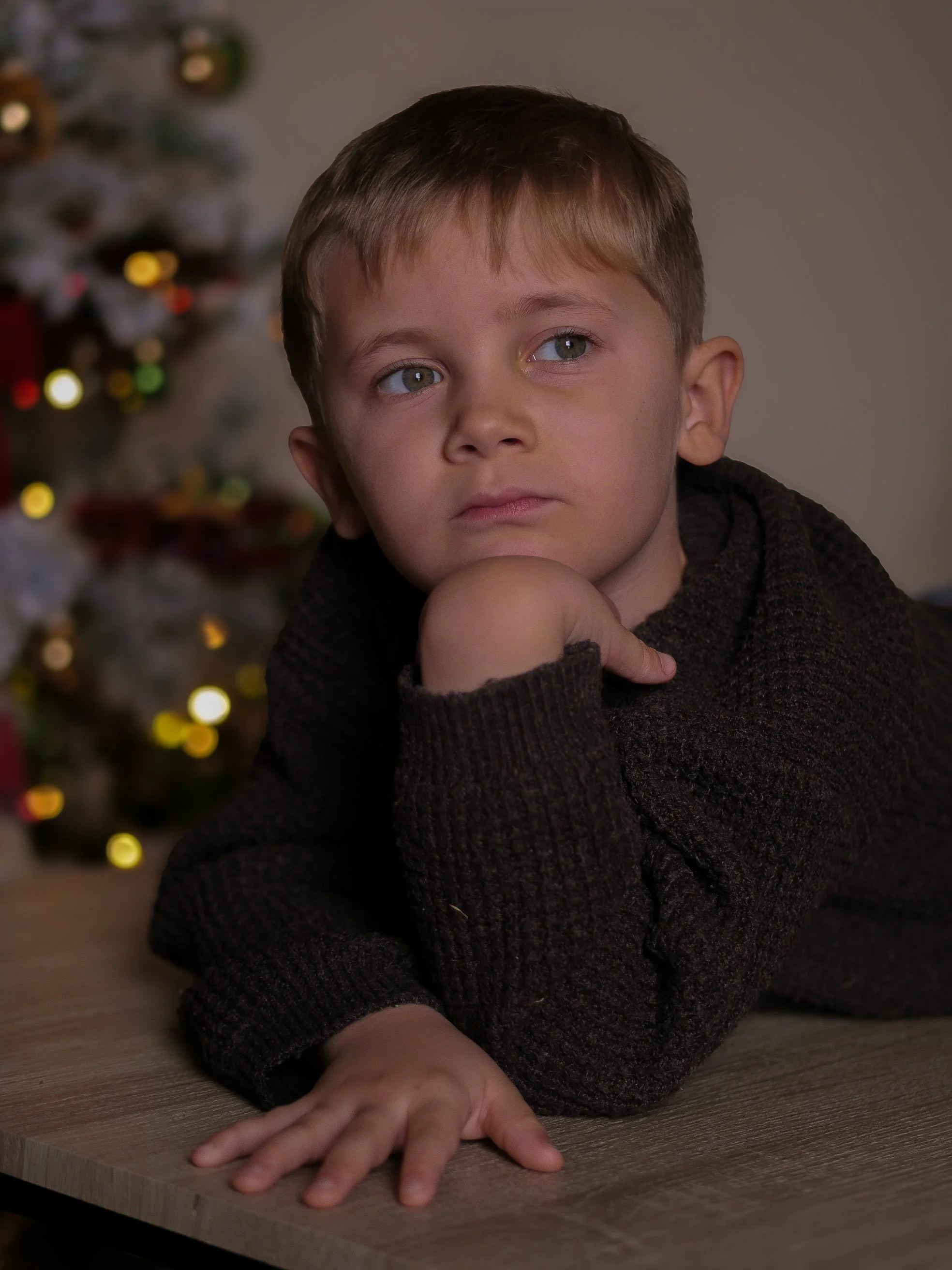 Jeune garçon blond avec des yeux bleus, assis à une table en bois, supportant sa tête avec sa main, avec un sapin de Noël décoré flou en arrière-plan.