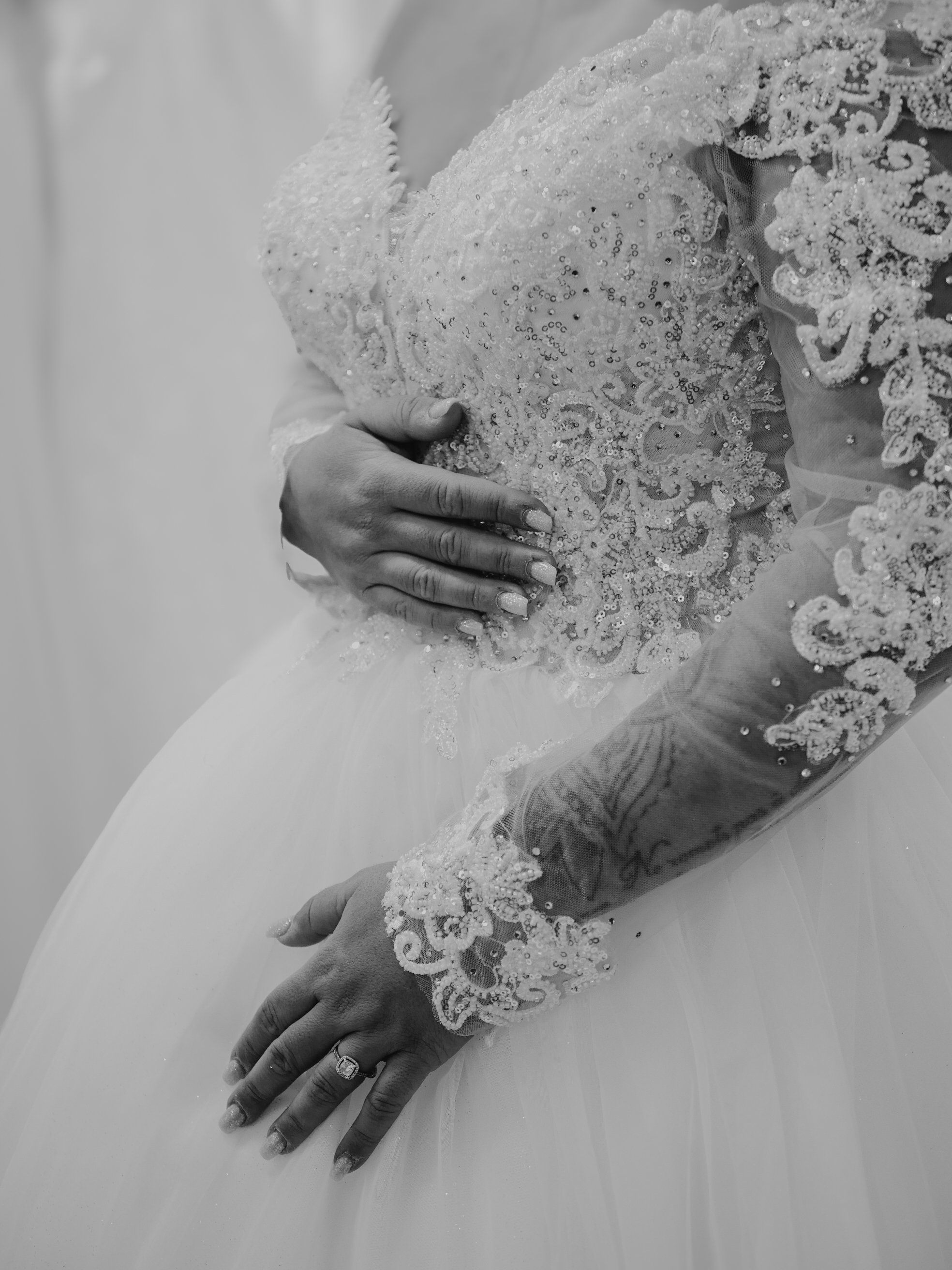 Une main d'une femme portant un anneau de mariage repose sur le ventre d'une personne portant une robe de mariée ornée de sequins et de broderies.